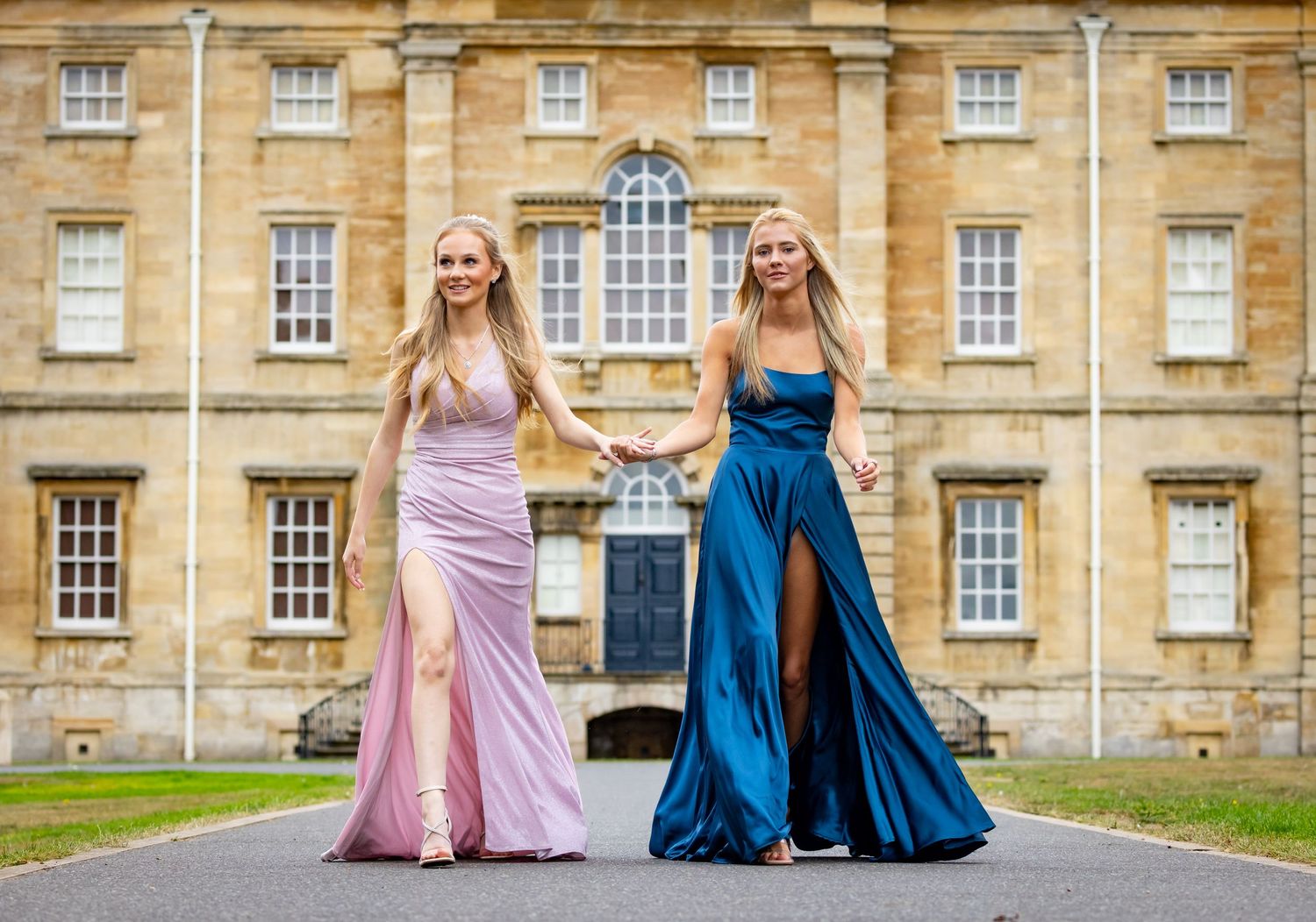Two people in formal evening gowns, one pink and one blue, stand together in front of a grand historic building.