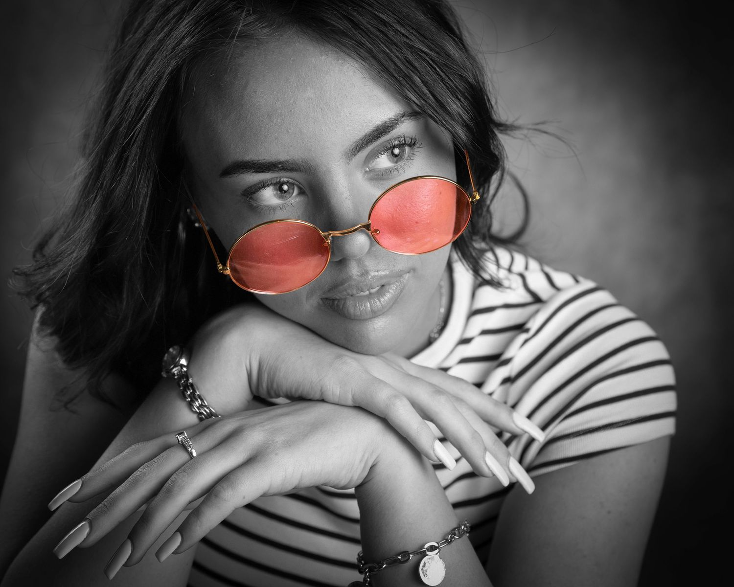 Black and white portrait of a young woman wearing red tinted oval sunglasses and a striped shirt, smiling warmly.