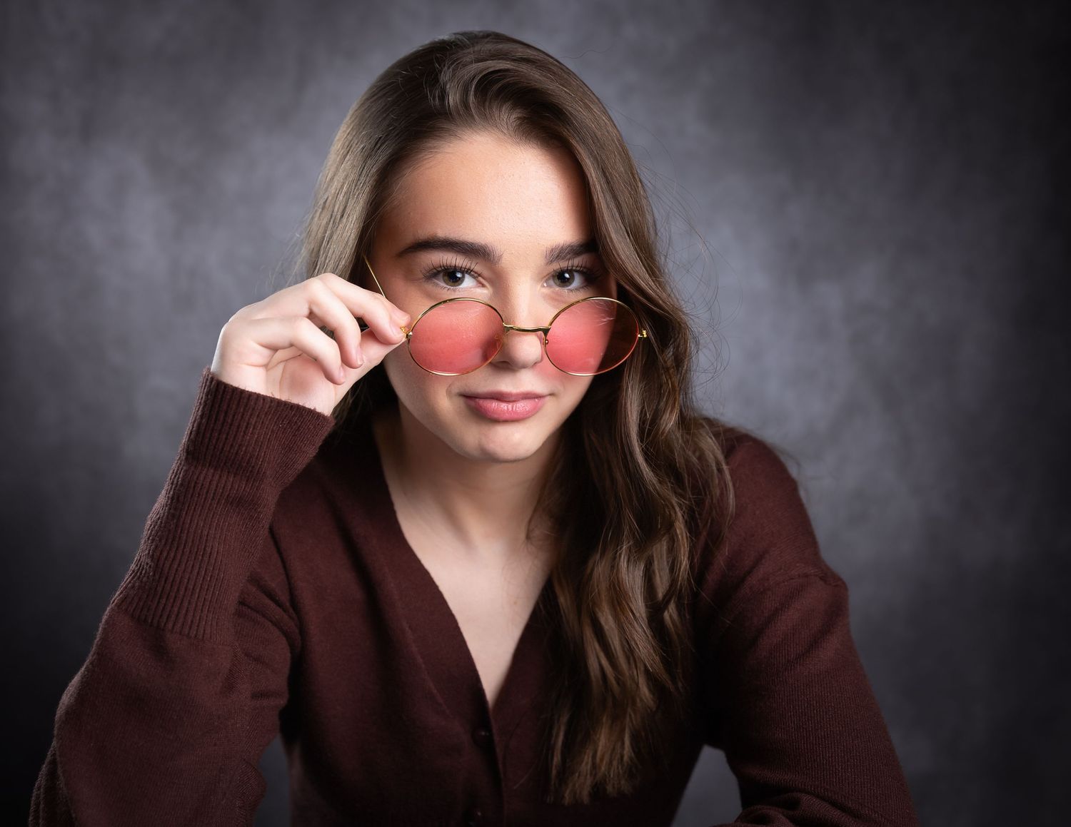Young woman with long brown hair holding pink round sunglasses in a studio portrait.