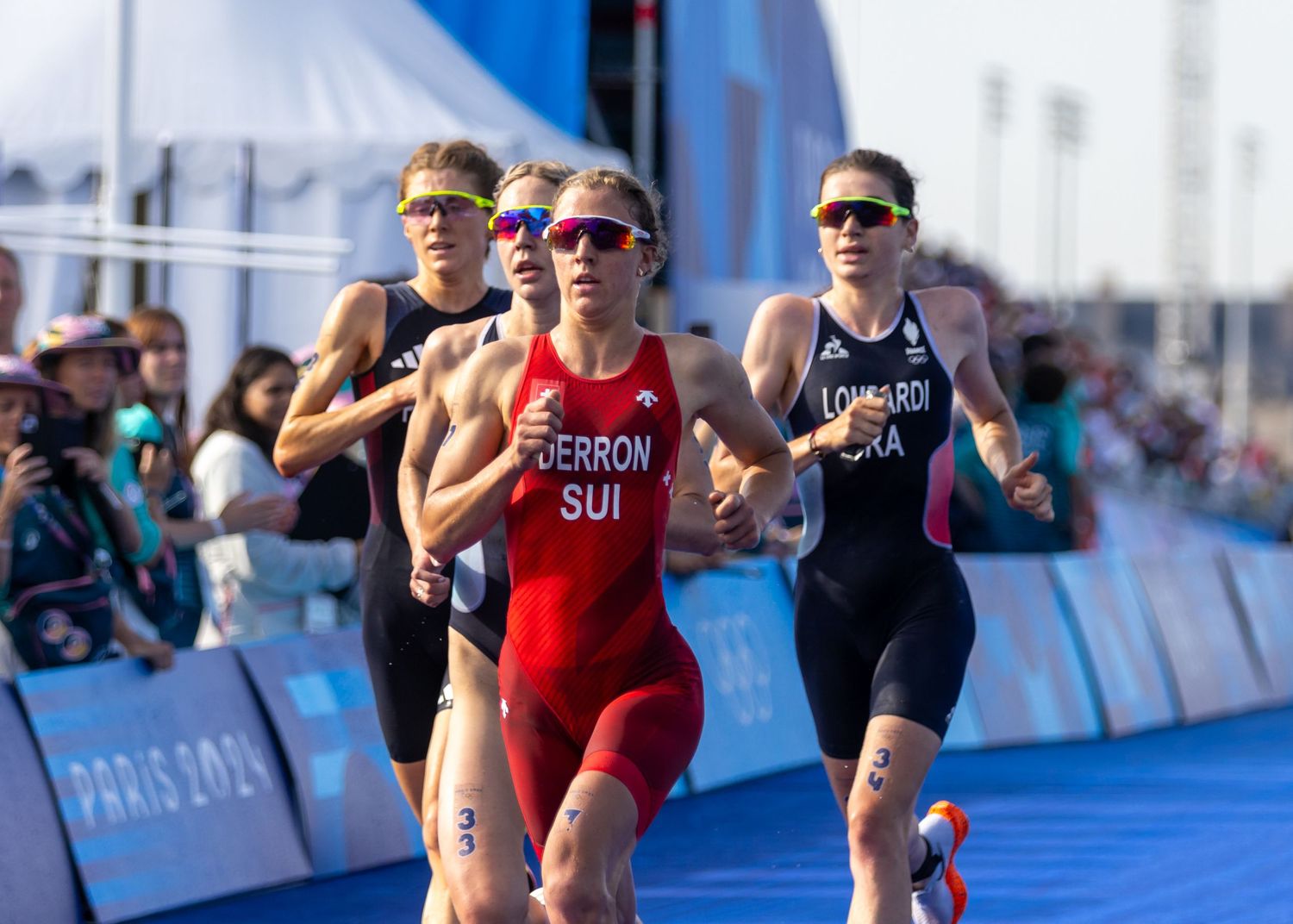 Athletes compete in a triathlon running event with crowd barriers and spectators.