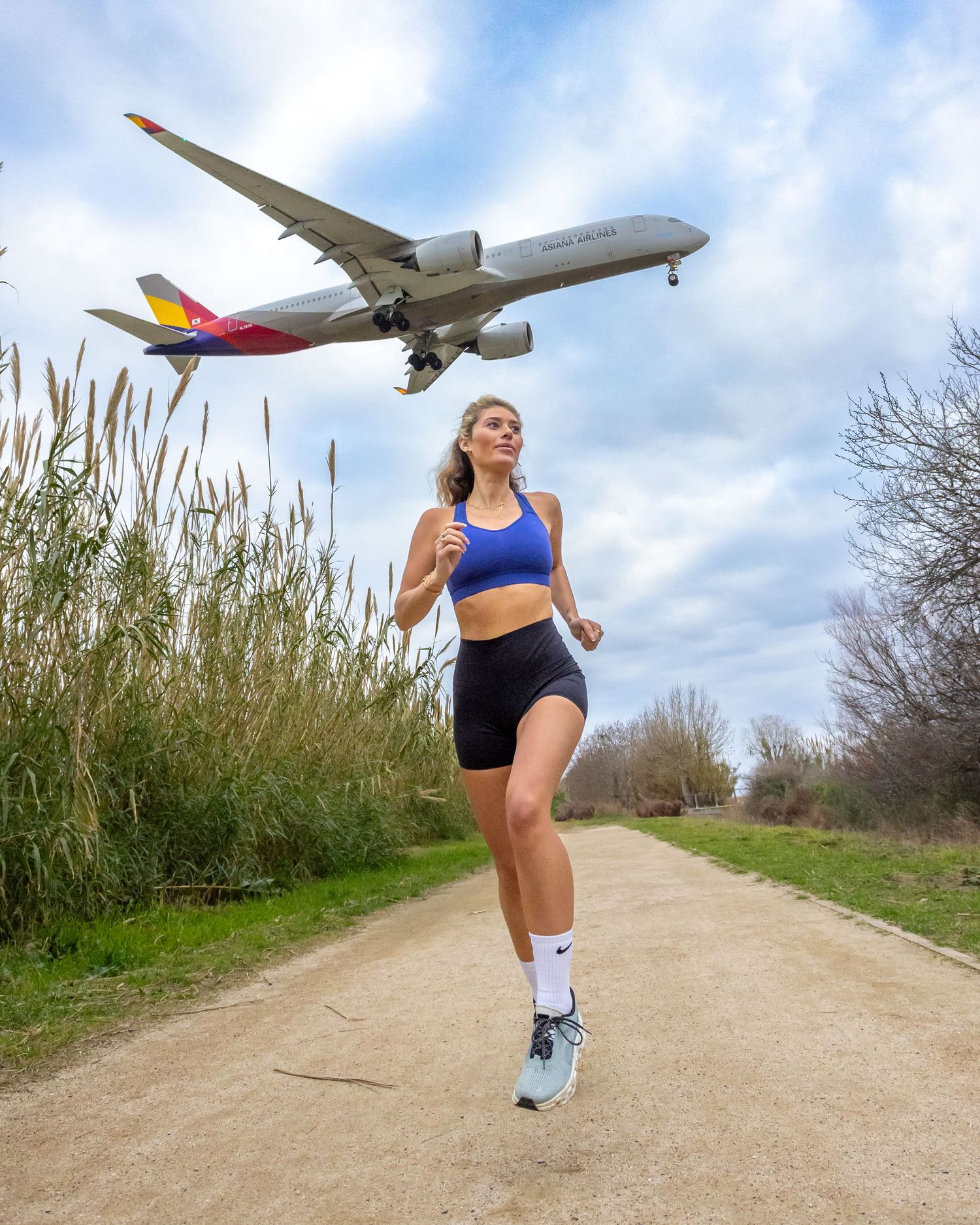 A commercial airliner soars overhead while someone exercises on a dirt path bordered by tall grass and shrubs.
