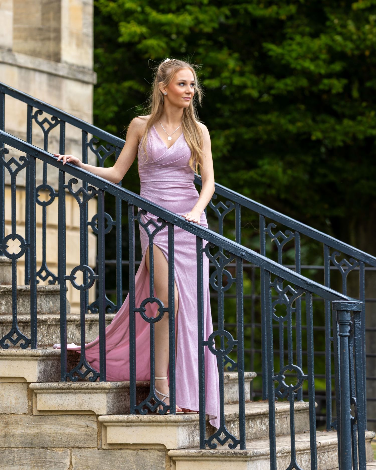 Person in a lavender dress poses elegantly on stone steps with ornate metal railings in an outdoor setting.