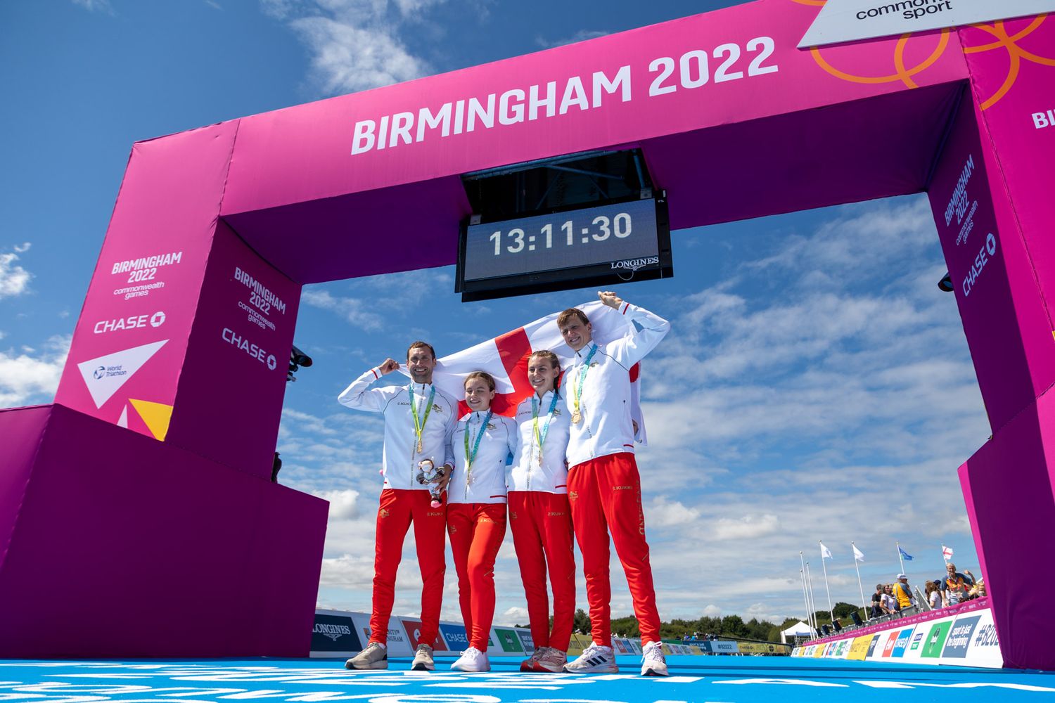 british Athletes celebrating under pink Birmingham 2022 finish arch against blue sky.