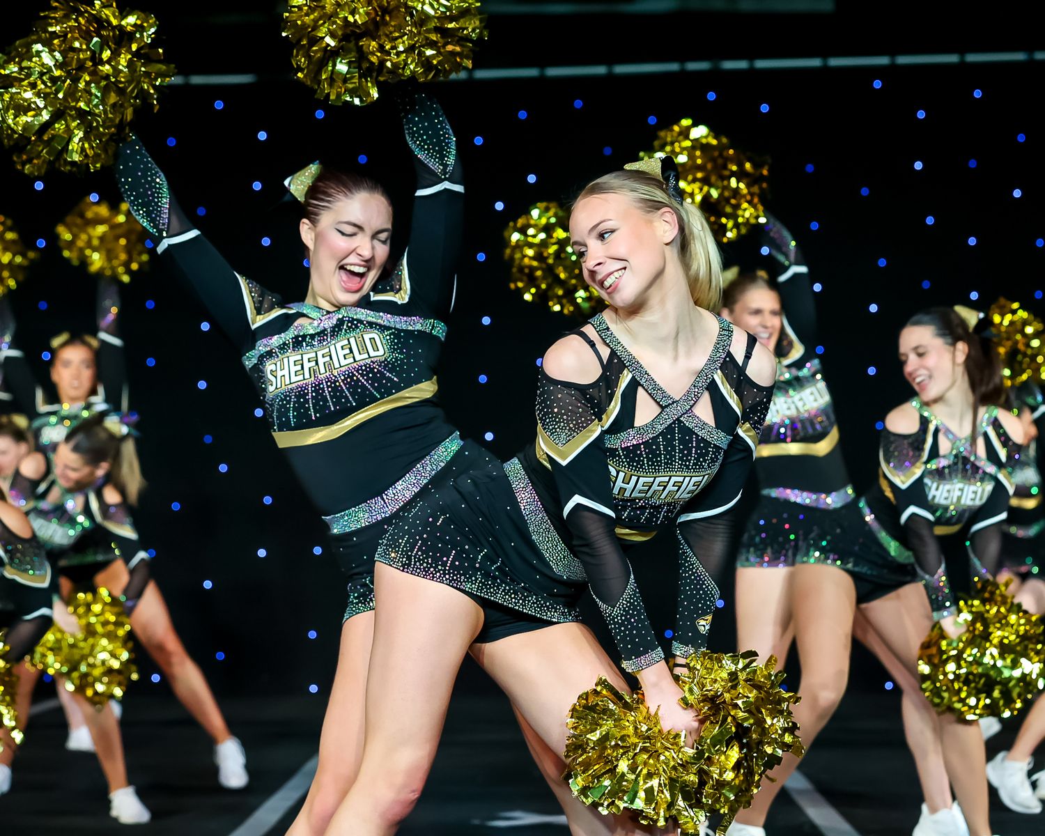 Cheerleaders in navy and silver uniforms perform an energetic routine with pom poms against a sparkling backdrop.