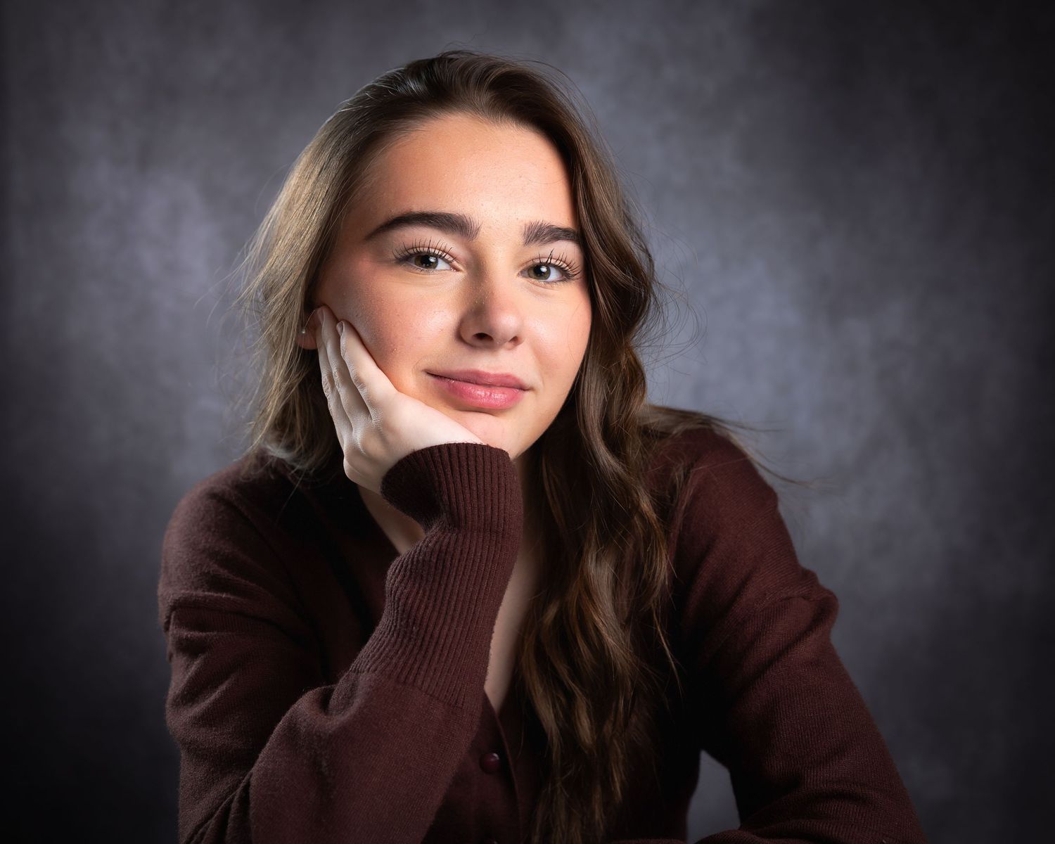 Young woman with long brown hair posing thoughtfully with hand on cheek against gray studio background.