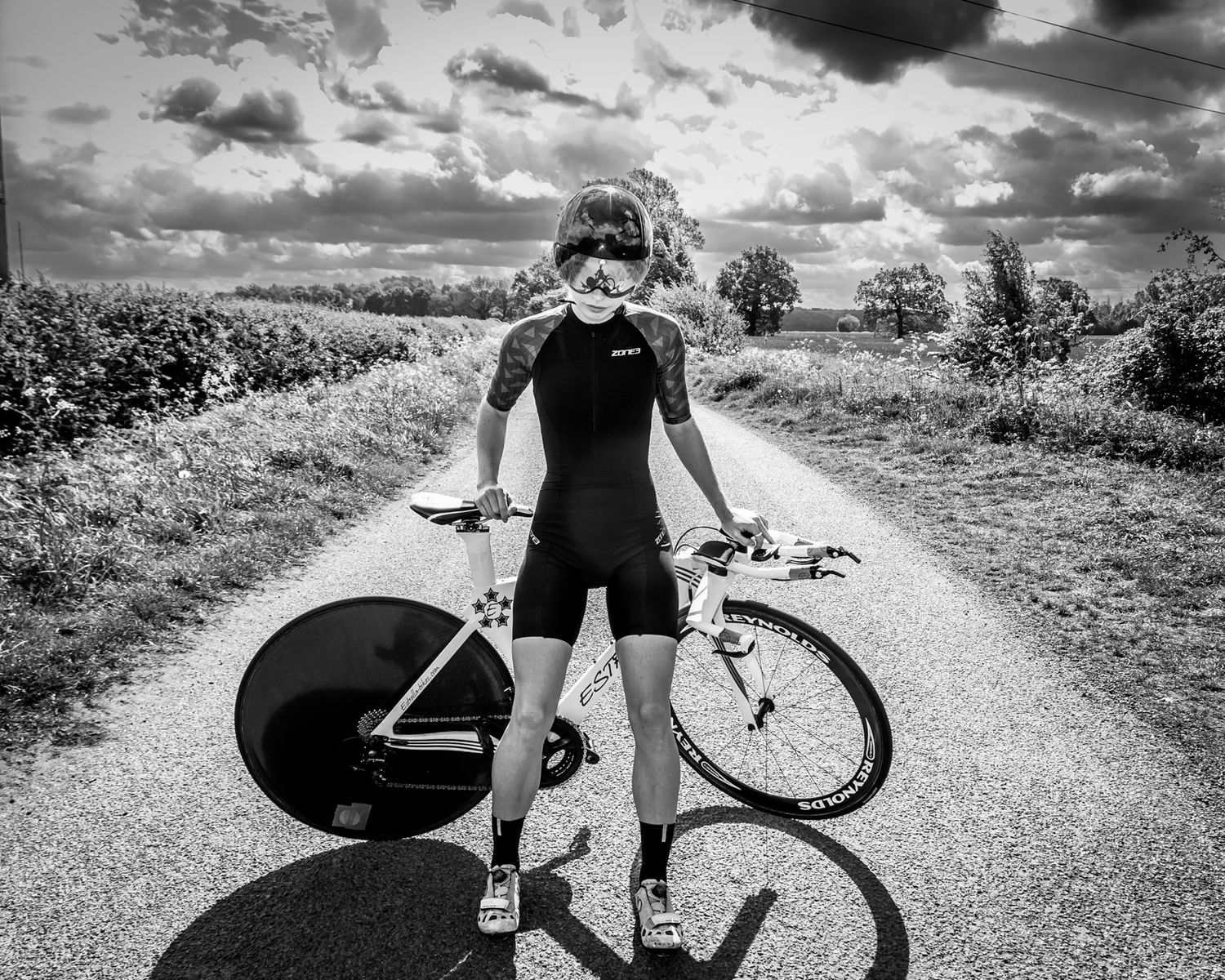 A cyclist in a black outfit stands on a rural road with a racing bike against a dramatic cloudy sky.
