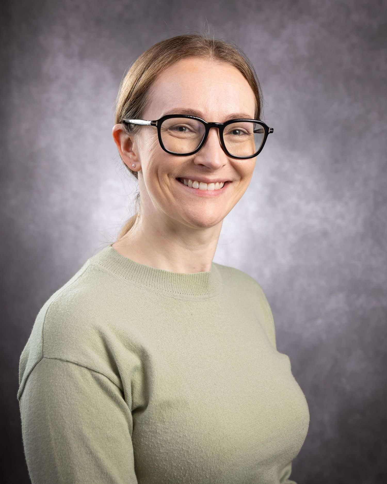 Professional headshot portrait of someone wearing glasses and a sage green sweater against a textured gray background.