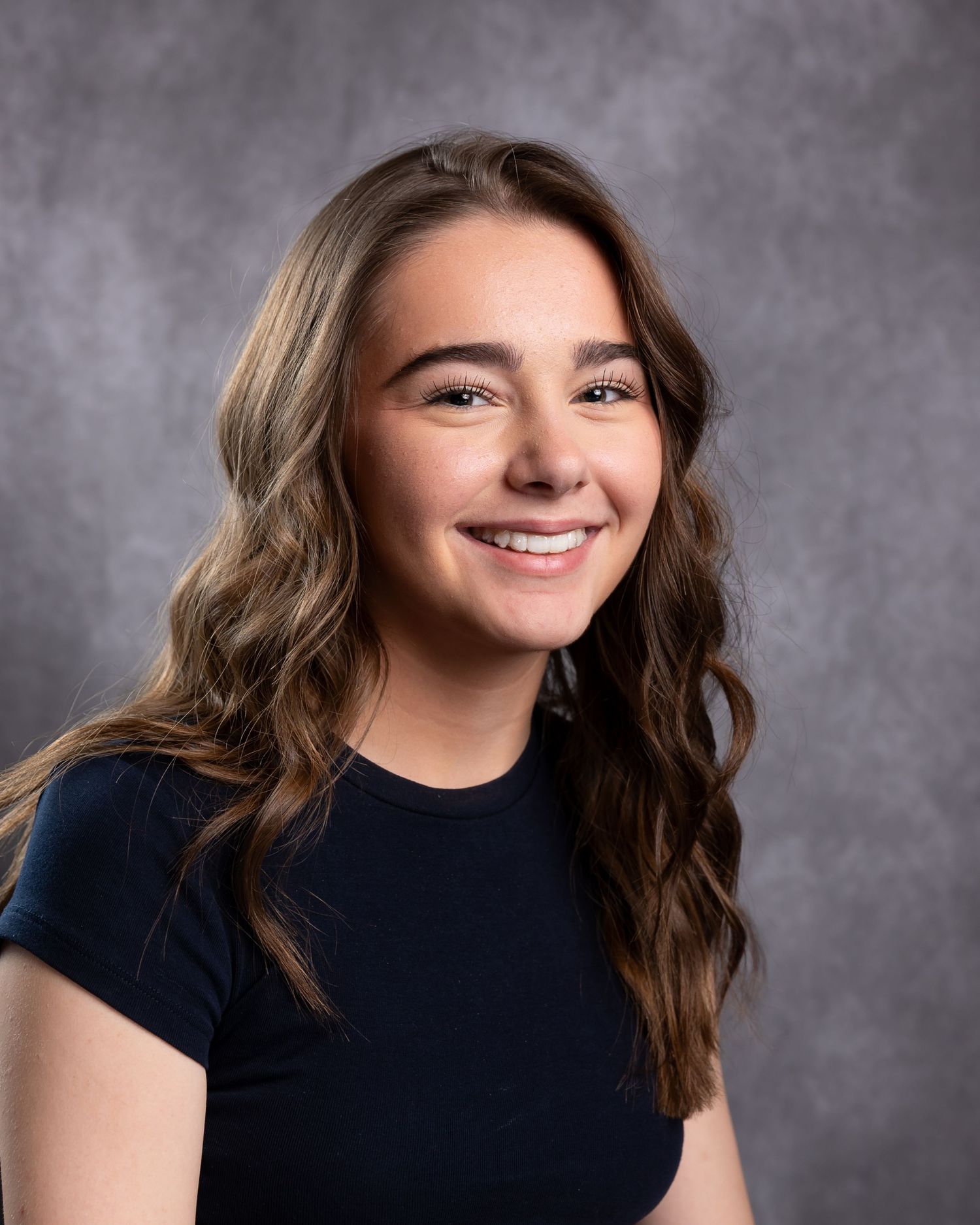 Smiling young woman with long wavy brown hair wearing a navy top against a gray studio background.