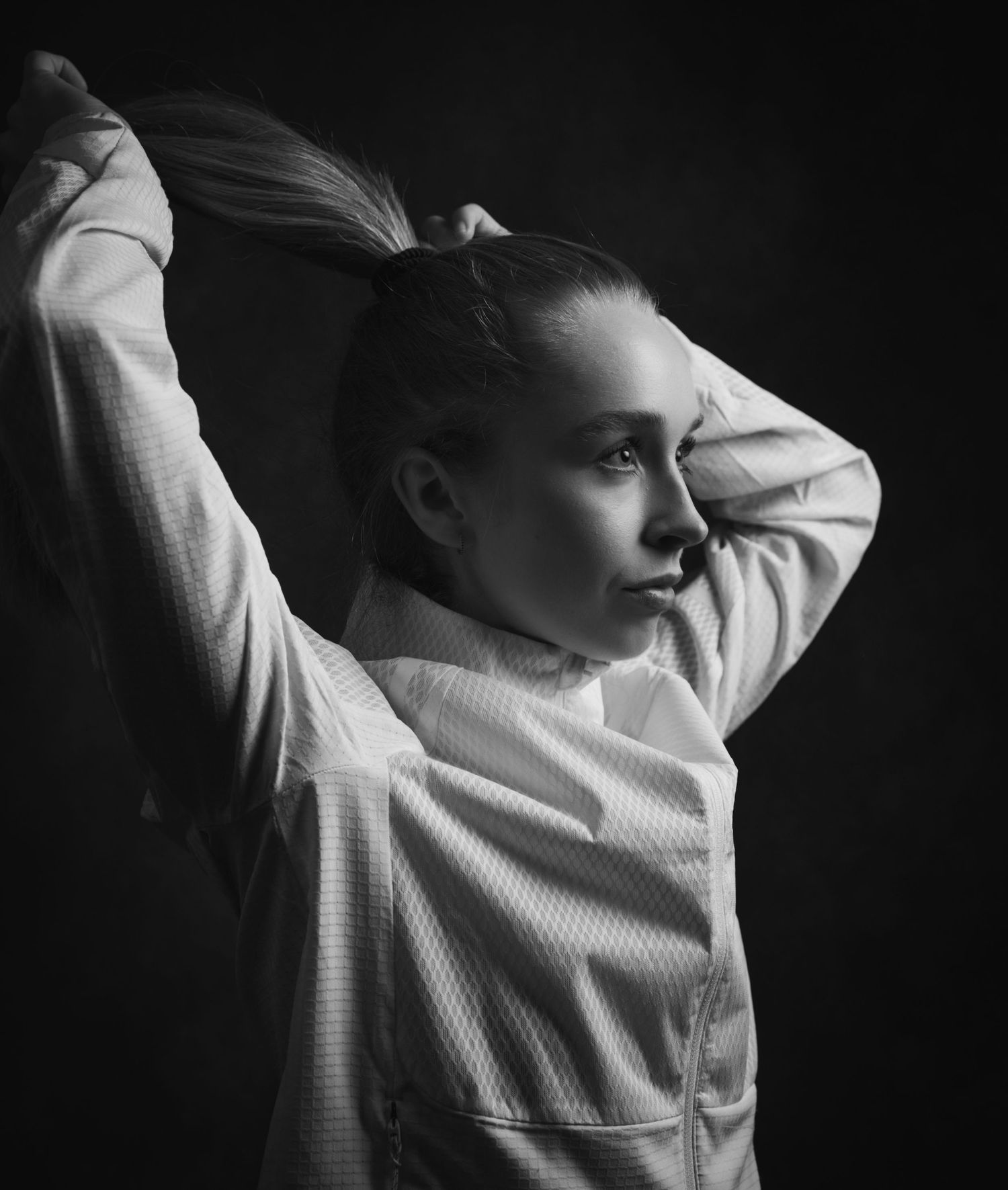 Black and white portrait of a young woman in a white jacket tying her hair up against a dark background.