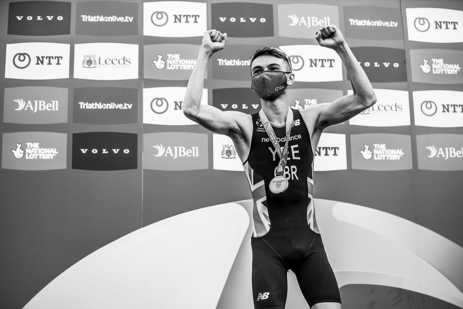 Black and white photo of a triathlete alex yee celebrating on podium with raised arms at competition with sponsor logos behind