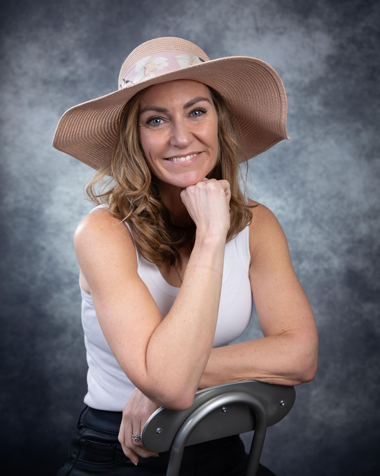 Studio portrait with wide brimmed tan hat and white tank top against dramatic gray backdrop.