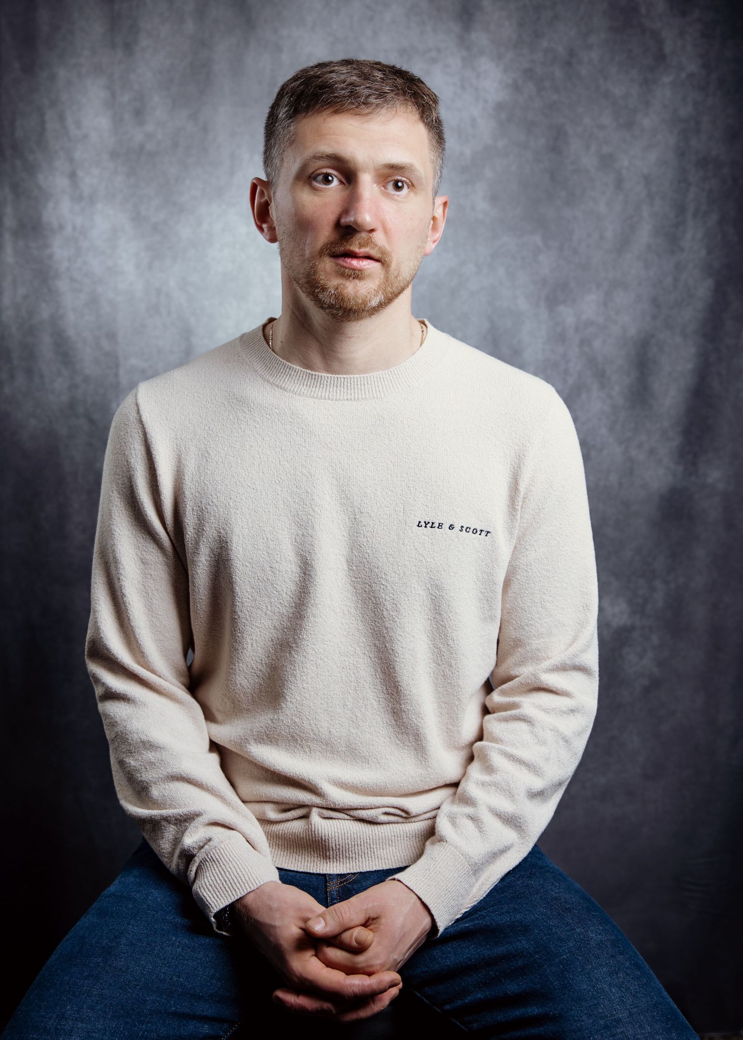 A person wearing a cream colored sweatshirt and blue jeans sits against a dark textured background.