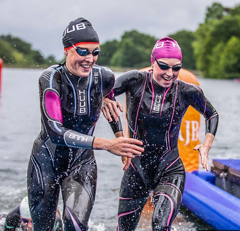 Two triathletes in wetsuits laugh together while emerging from the water during a race.