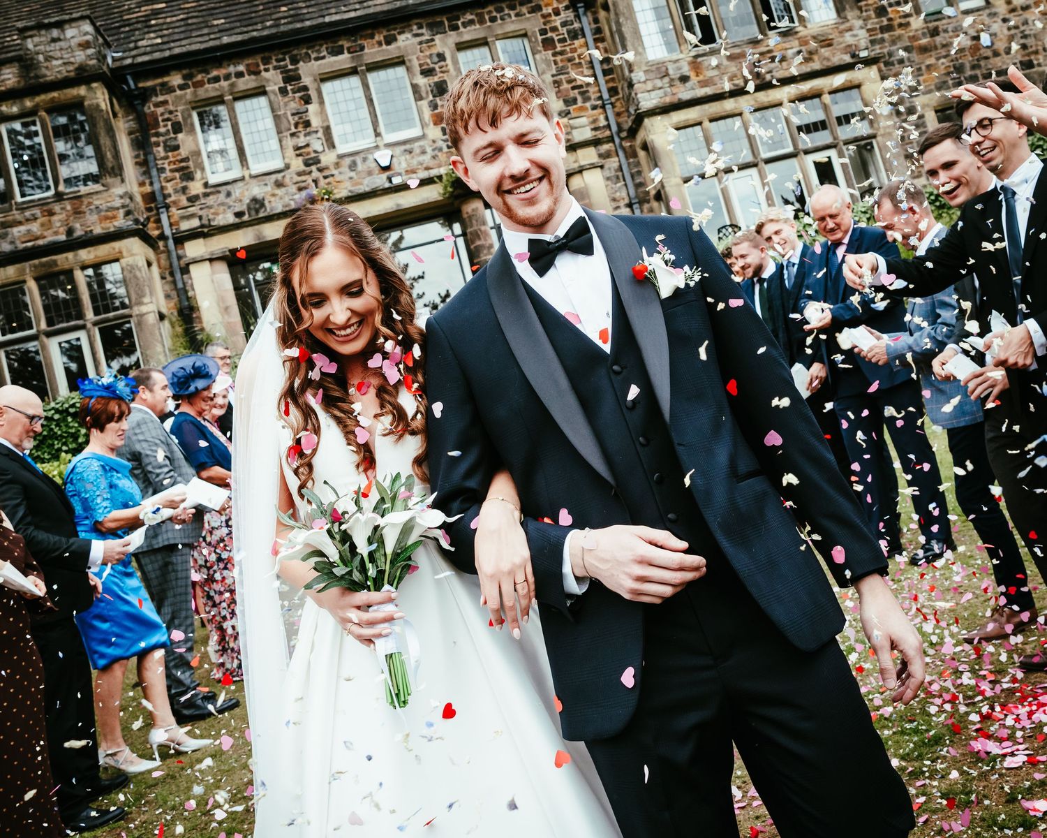 Newlyweds celebrate joyfully amid shower of confetti outside historic stone building during wedding ceremony.