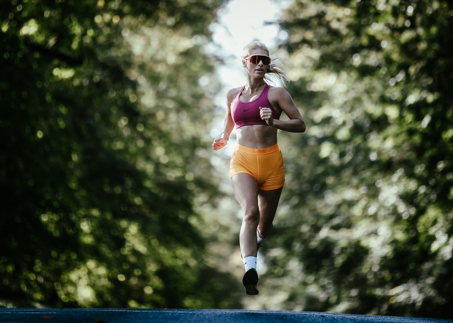 Woman in pink sports bra and orange shorts running outdoors on a tree-lined path on a sunny day in a national park.