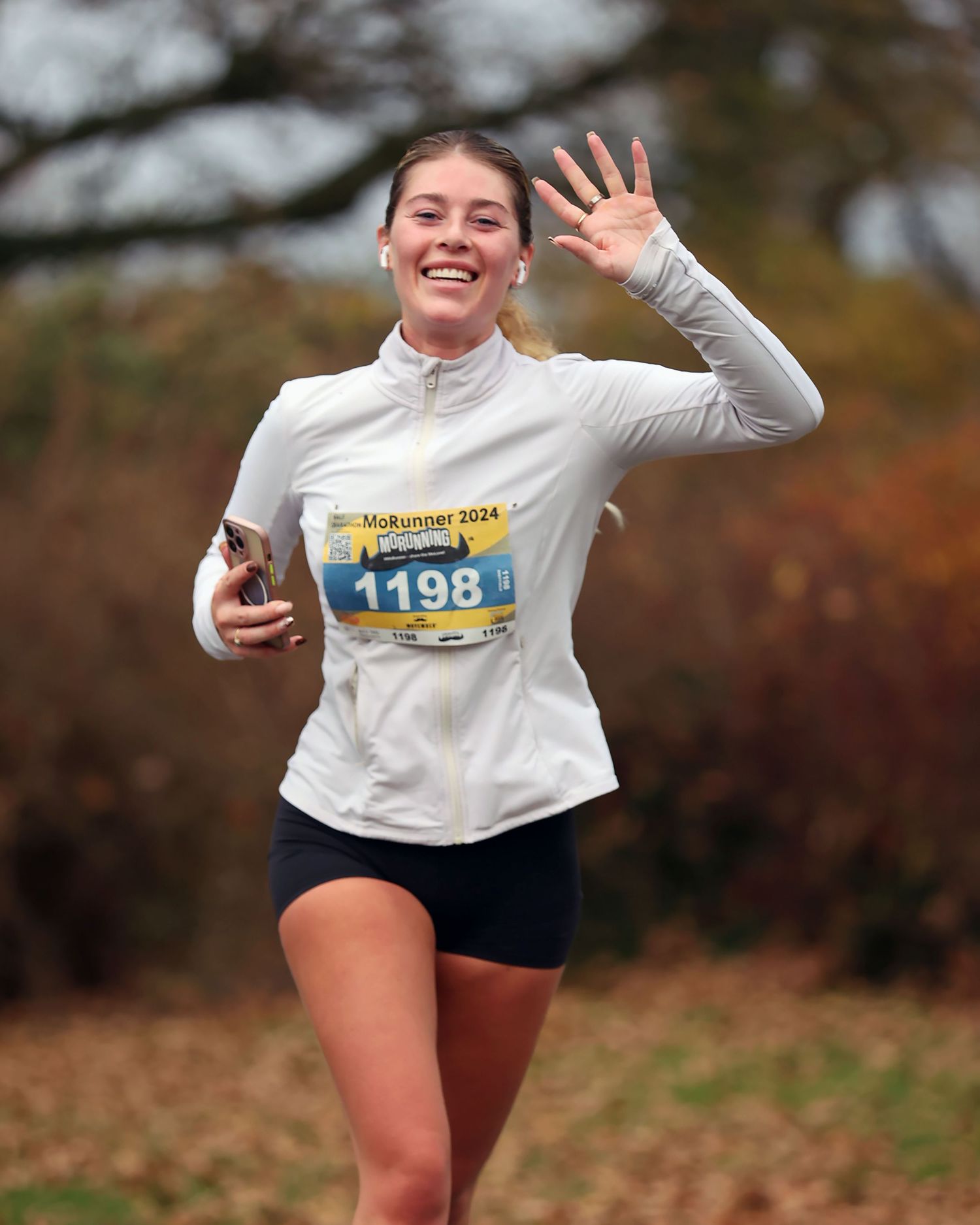 Runner in white top and race bib waving while participating in outdoor race.