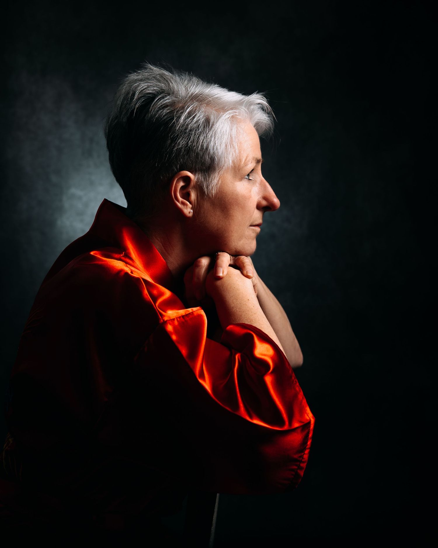 Dramatic side profile portrait against dark background with person in bright red garment with hand thoughtfully at chin.