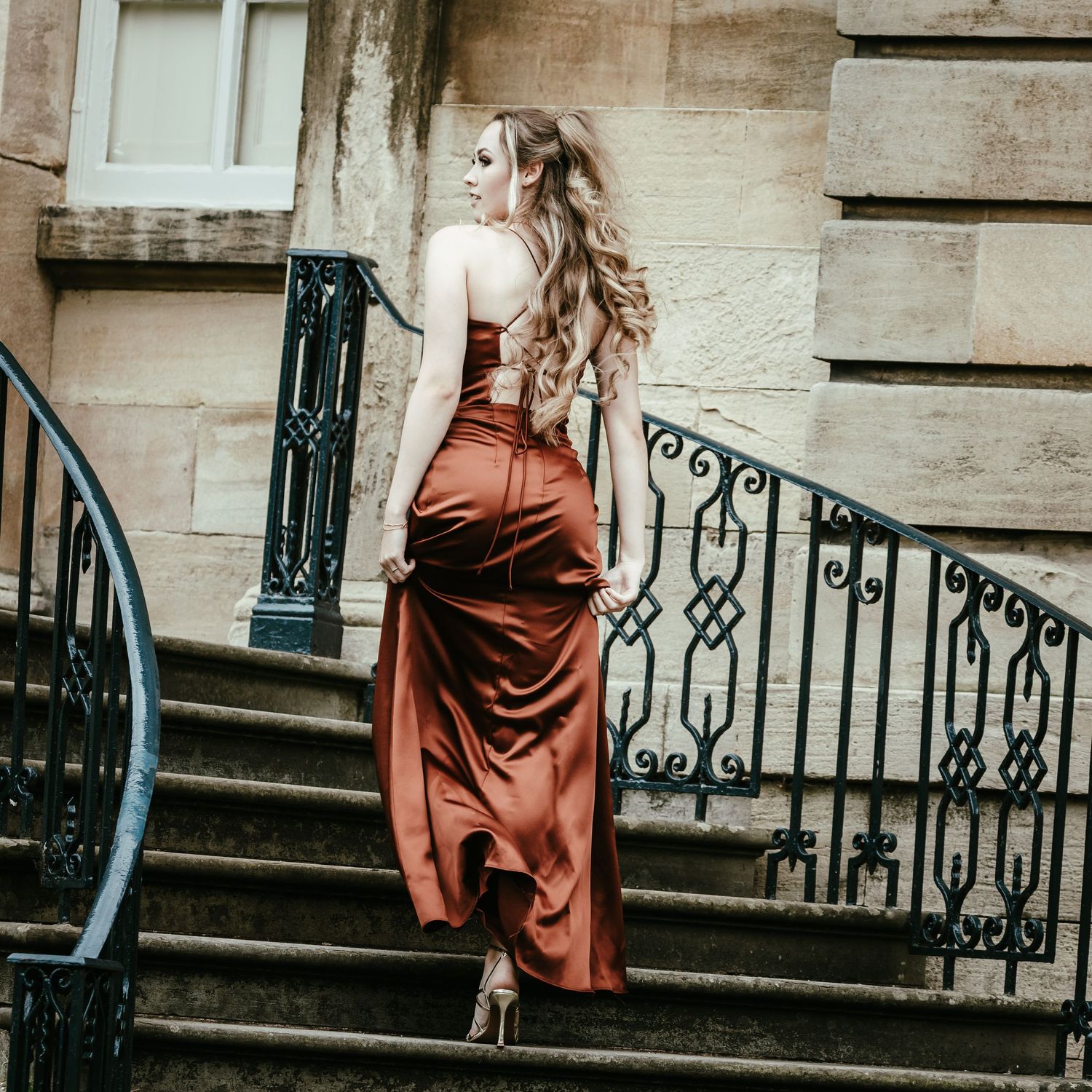 A long-haired figure in a rustic brown dress stands on vintage stone steps with ornate iron railings.