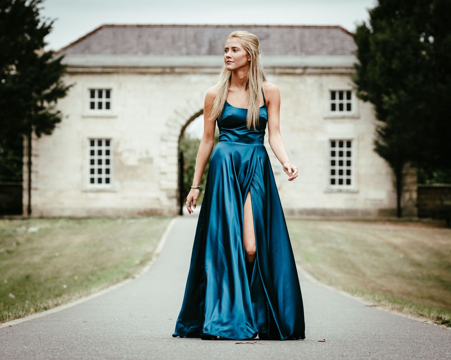 A stunning teal satin evening gown with a thigh-high slit stands out against a classic mansion backdrop.