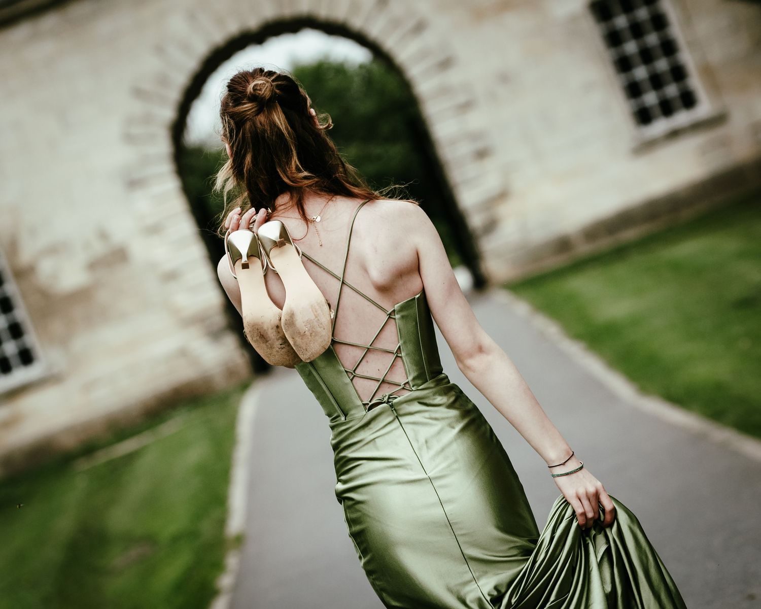 Woman in elegant green satin dress with criss-cross back detail poses outdoors near stone building.