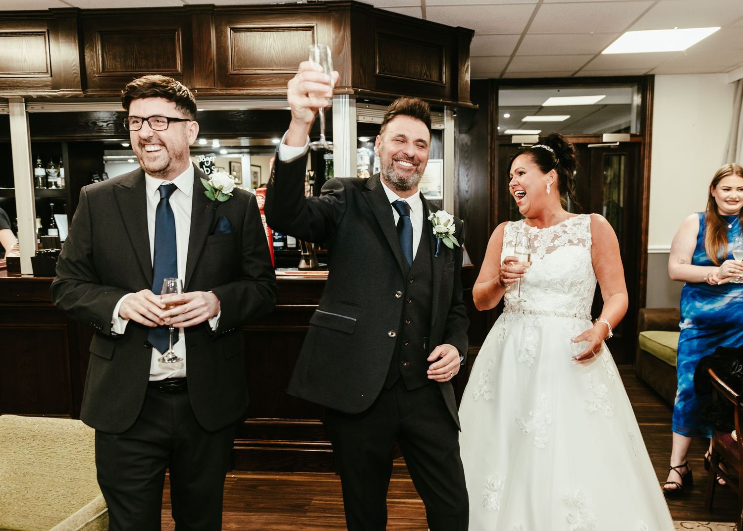 Wedding party members in formal attire laugh and share drinks in a reception venue hallway.