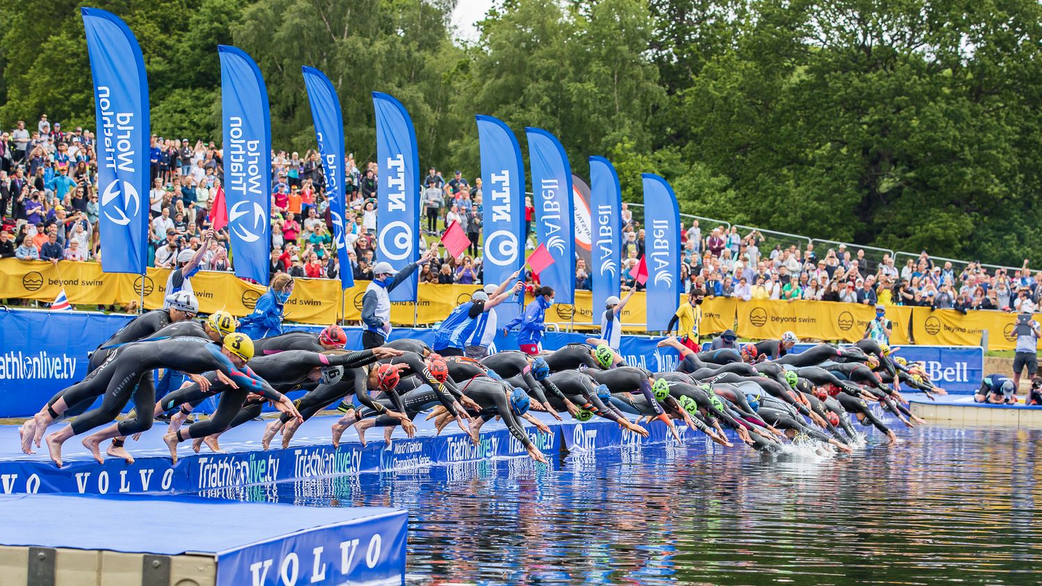 Athletes dive into the water at the start of a triathlon competition with blue sponsor banners and spectators in background.
