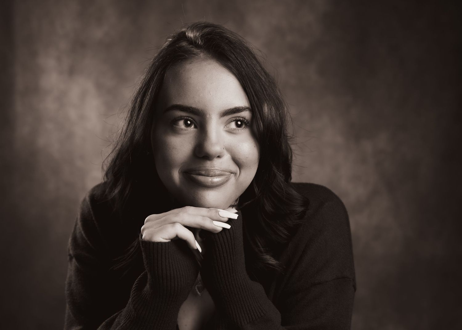 Black and white portrait of a young woman with long dark hair, smiling warmly with hands clasped under chin.