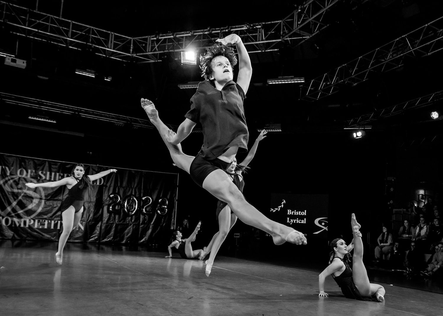 A dramatic black and white performance capture of a dancer leaping through the air on stage.
