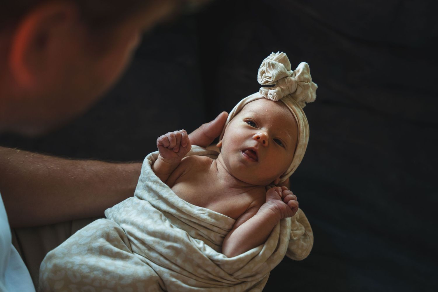 Close-up of newborn wrapped in a blanket with a bow, looking up with curiosity. / Newborn gewikkeld in een dekentje met strik, kijkt nieu