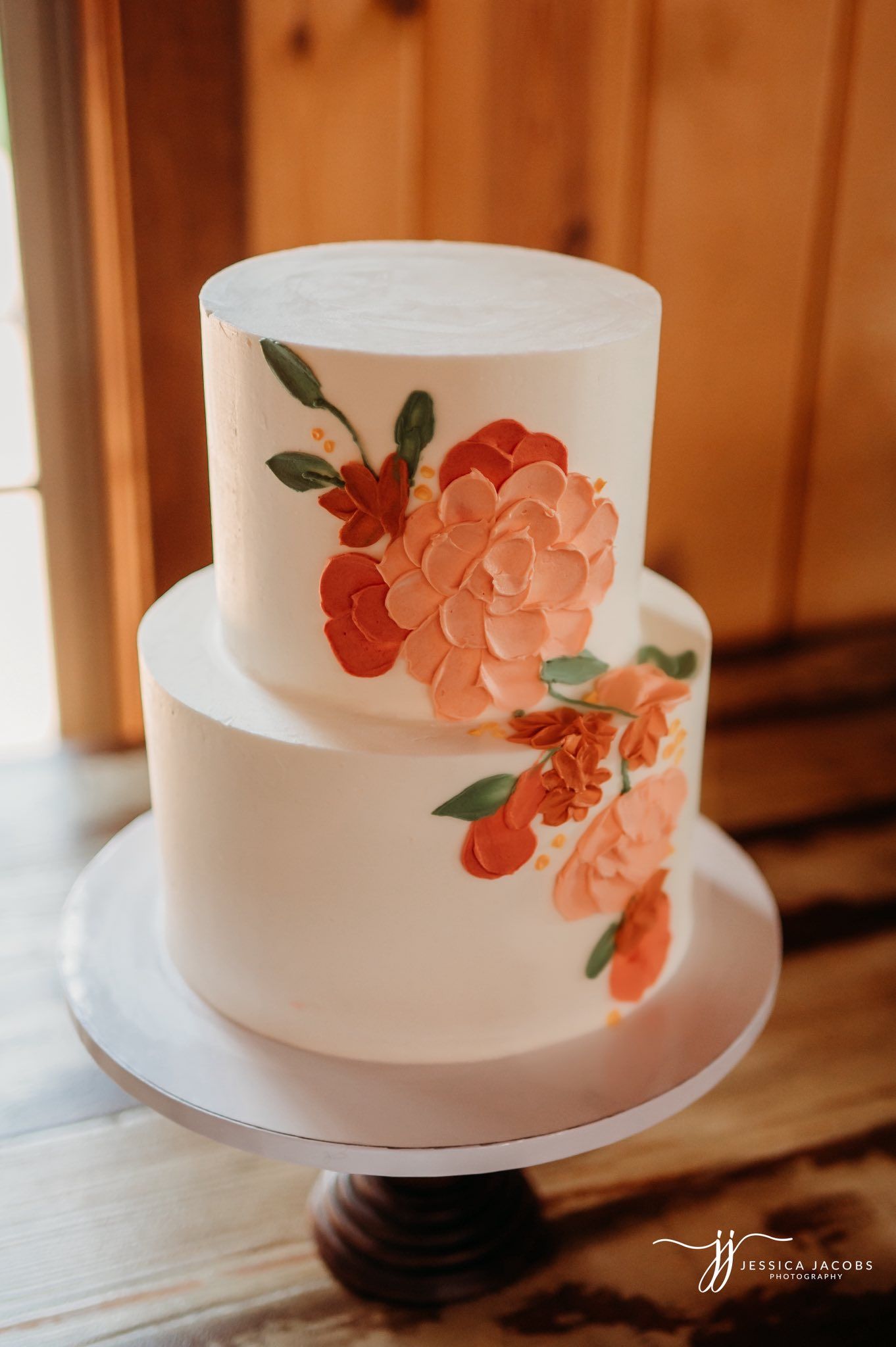 White wedding cake decorated with delicate coral-colored sugar flowers and green leaves displayed on a wooden surface.