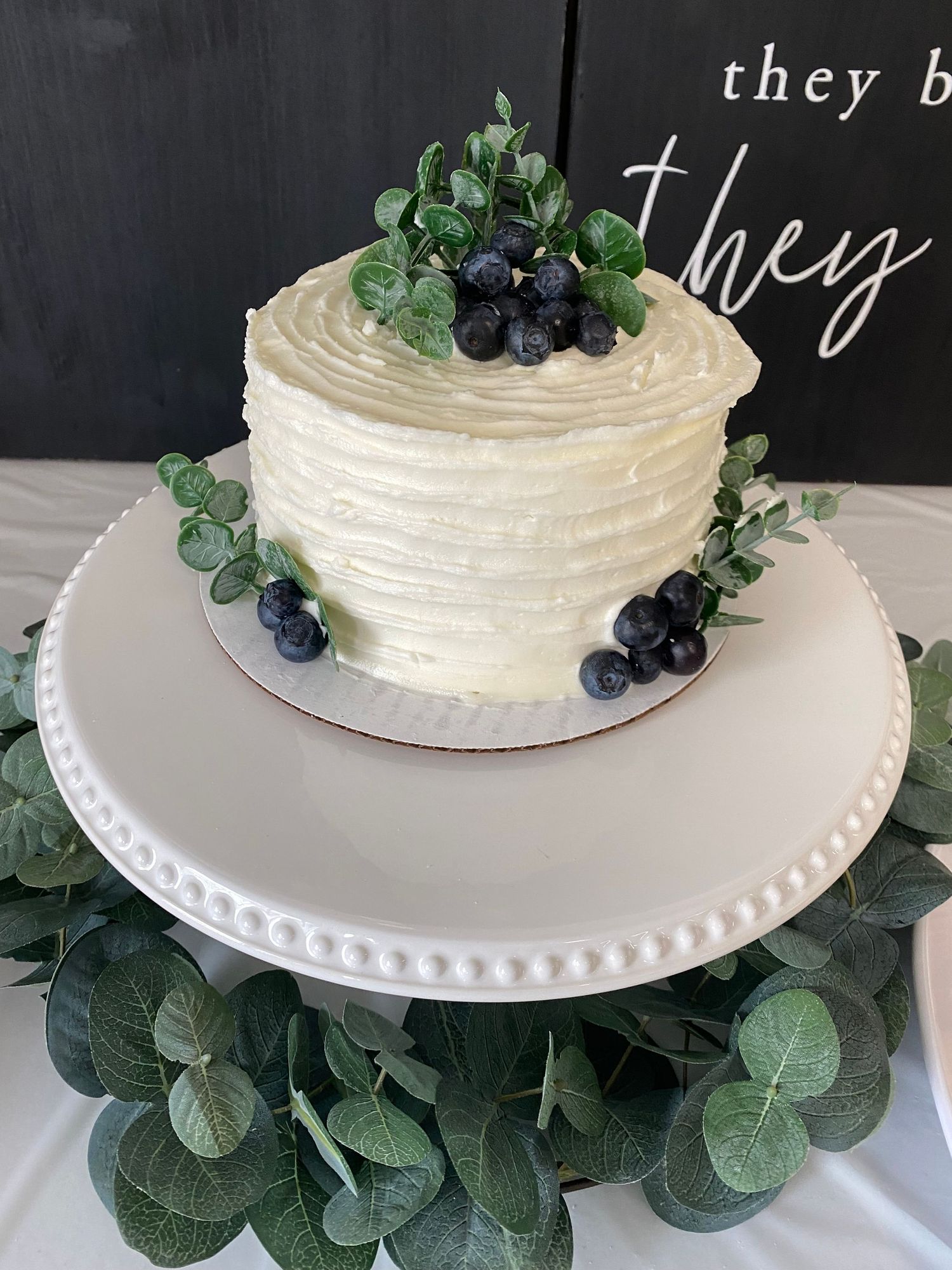 Single tier white wedding cake adorned with fresh blueberries and eucalyptus leaves on a white plate.