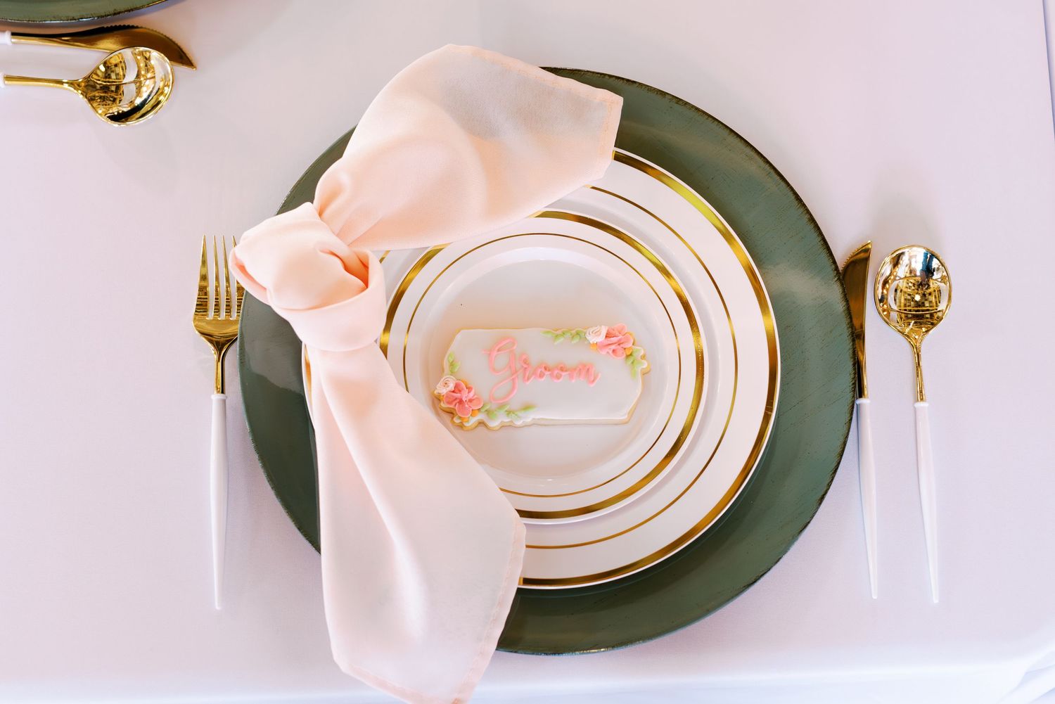 Elegant place setting with gold flatware, green charger plate, and pink napkin on white tablecloth.
