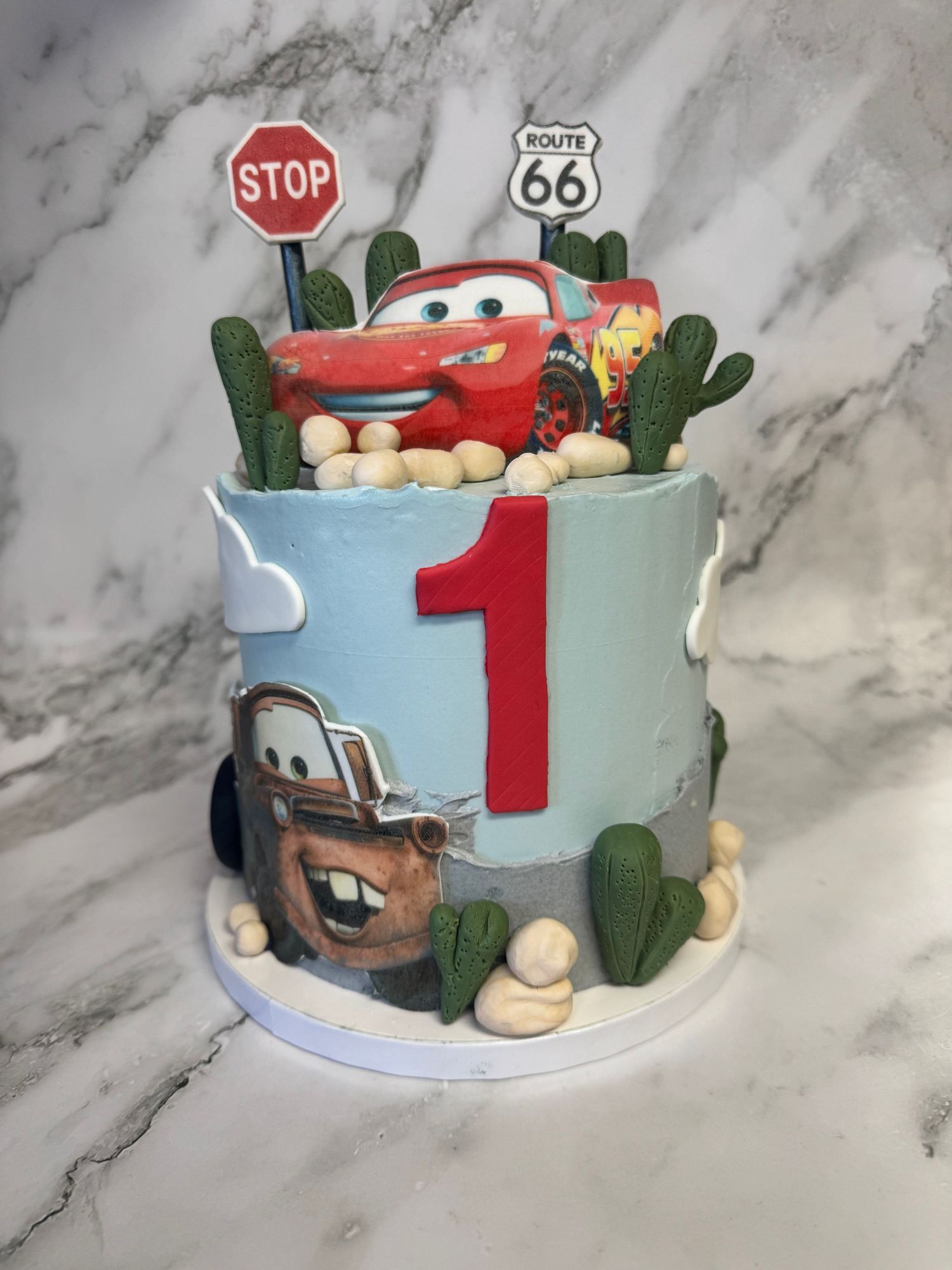 A light blue birthday cake decorated with Cars movie theme featuring Route 66 signs, cacti, and a red number one topper.