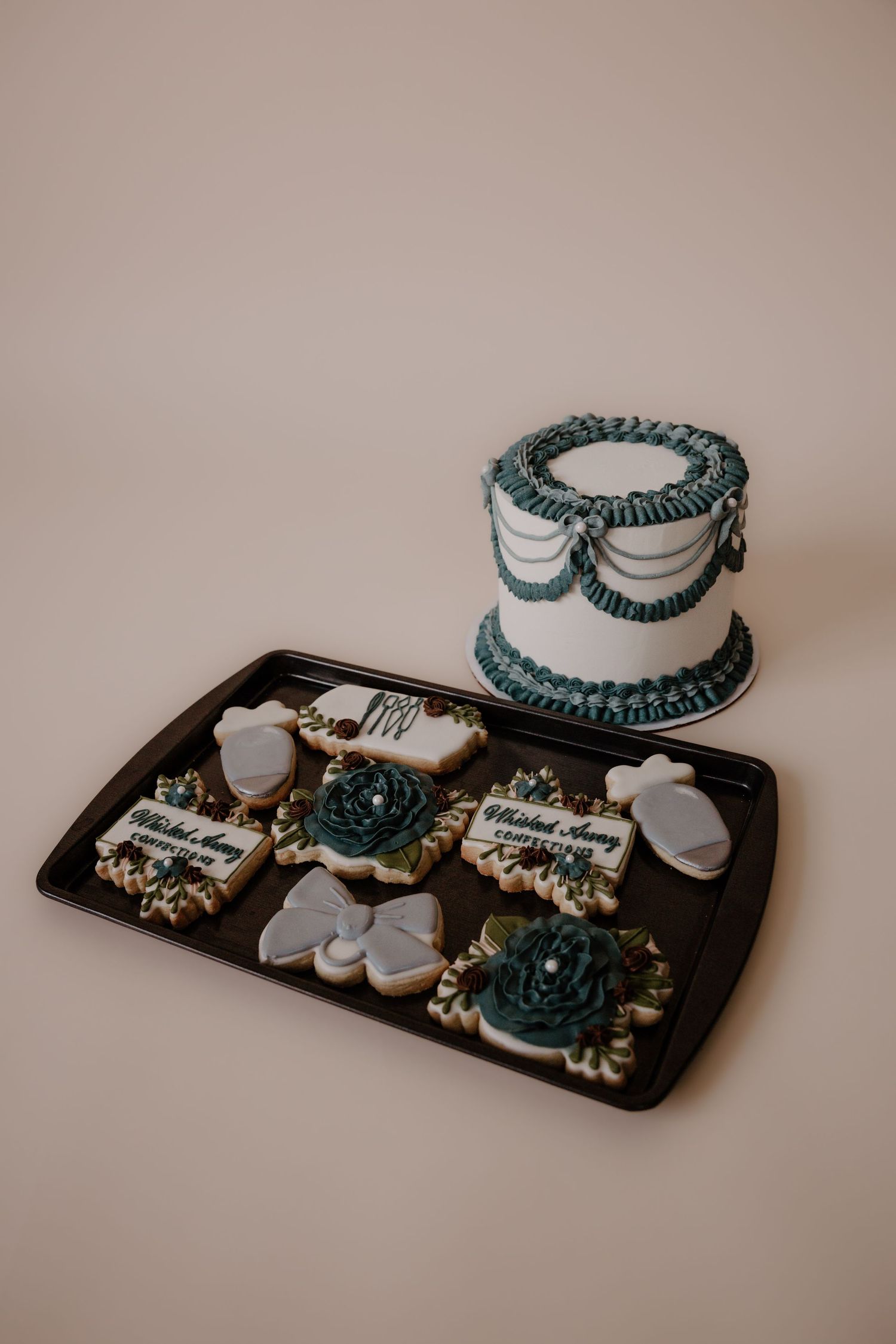 Vintage-style wedding cake with teal ruffle details alongside decorated cookies on dark platter.