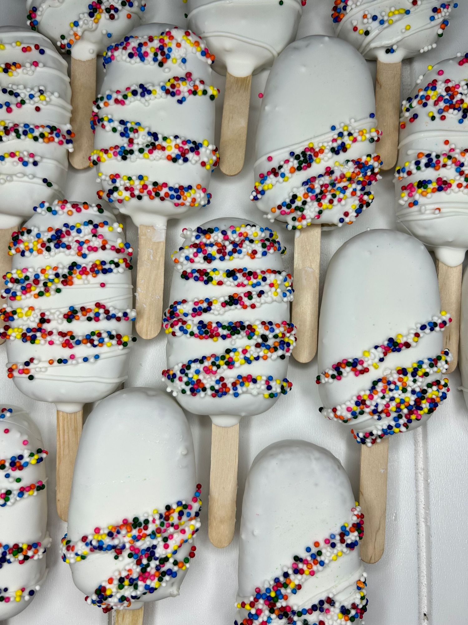White chocolate-dipped popsicles decorated with colorful sprinkles arranged in neat rows.