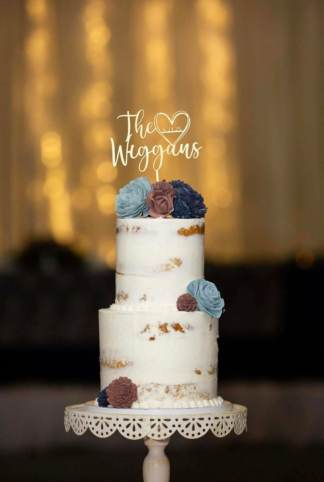 White wedding cake with blue and burgundy sugar flowers displayed on elegant white cake stand against golden bokeh background.