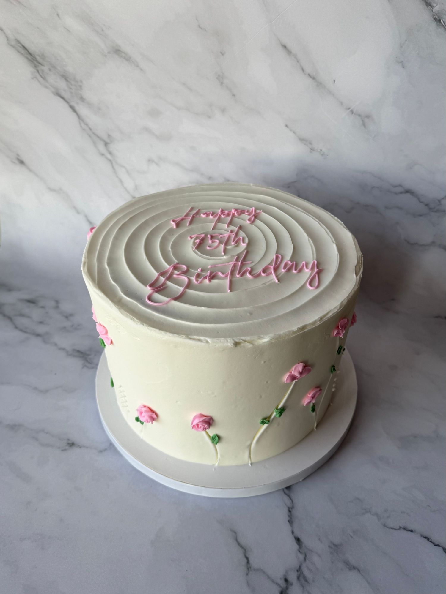 White buttercream birthday cake decorated with delicate pink flowers and spiral design on top, set on marble surface.