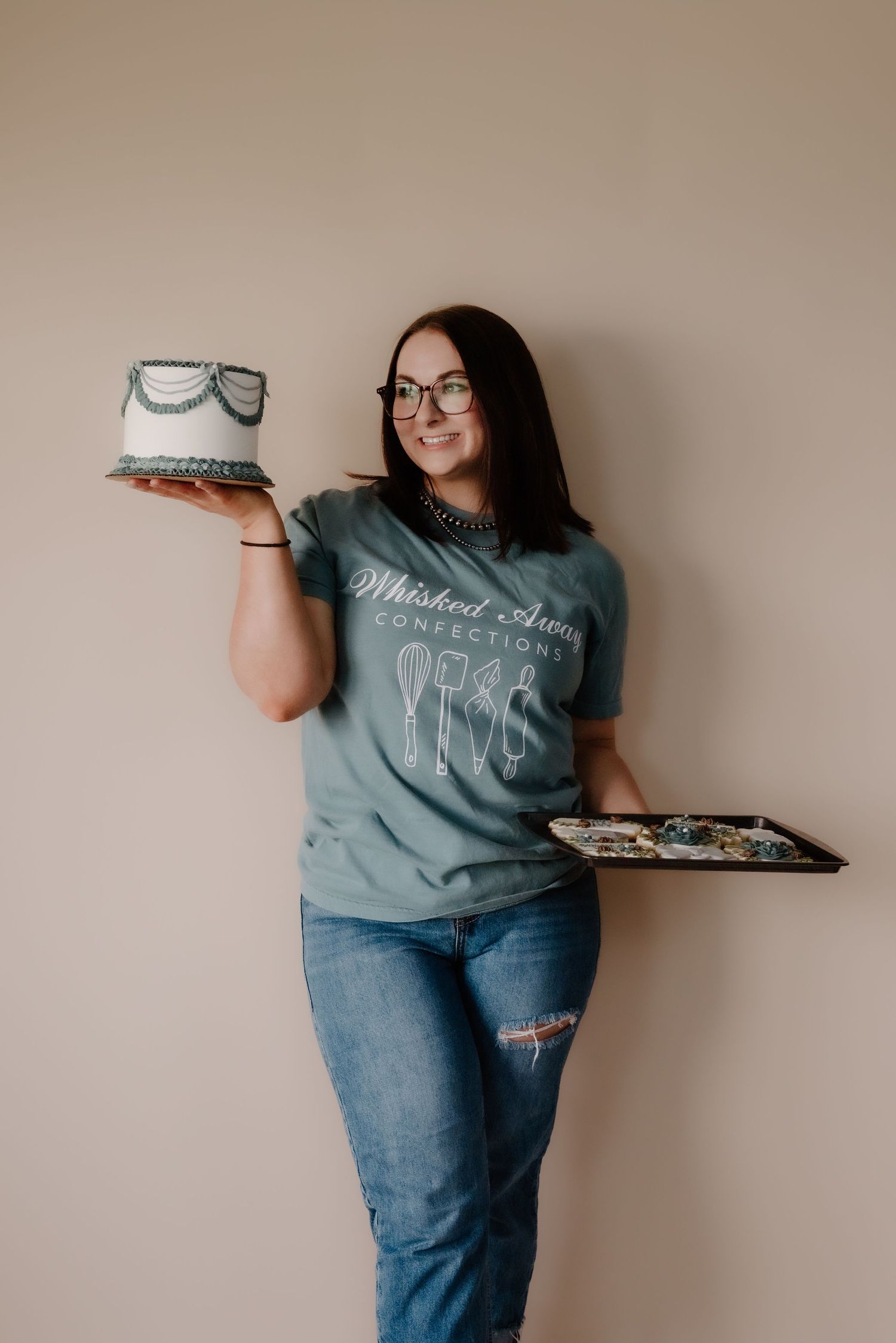 A person in a blue t-shirt and jeans poses with a cake against a beige wall.