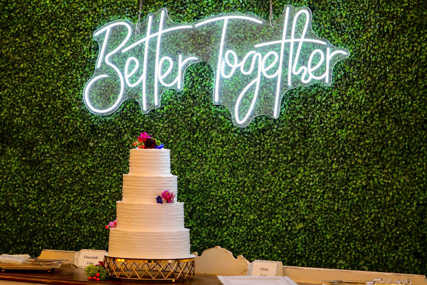 Wedding cake display with neon sign and greenery wall backdrop.