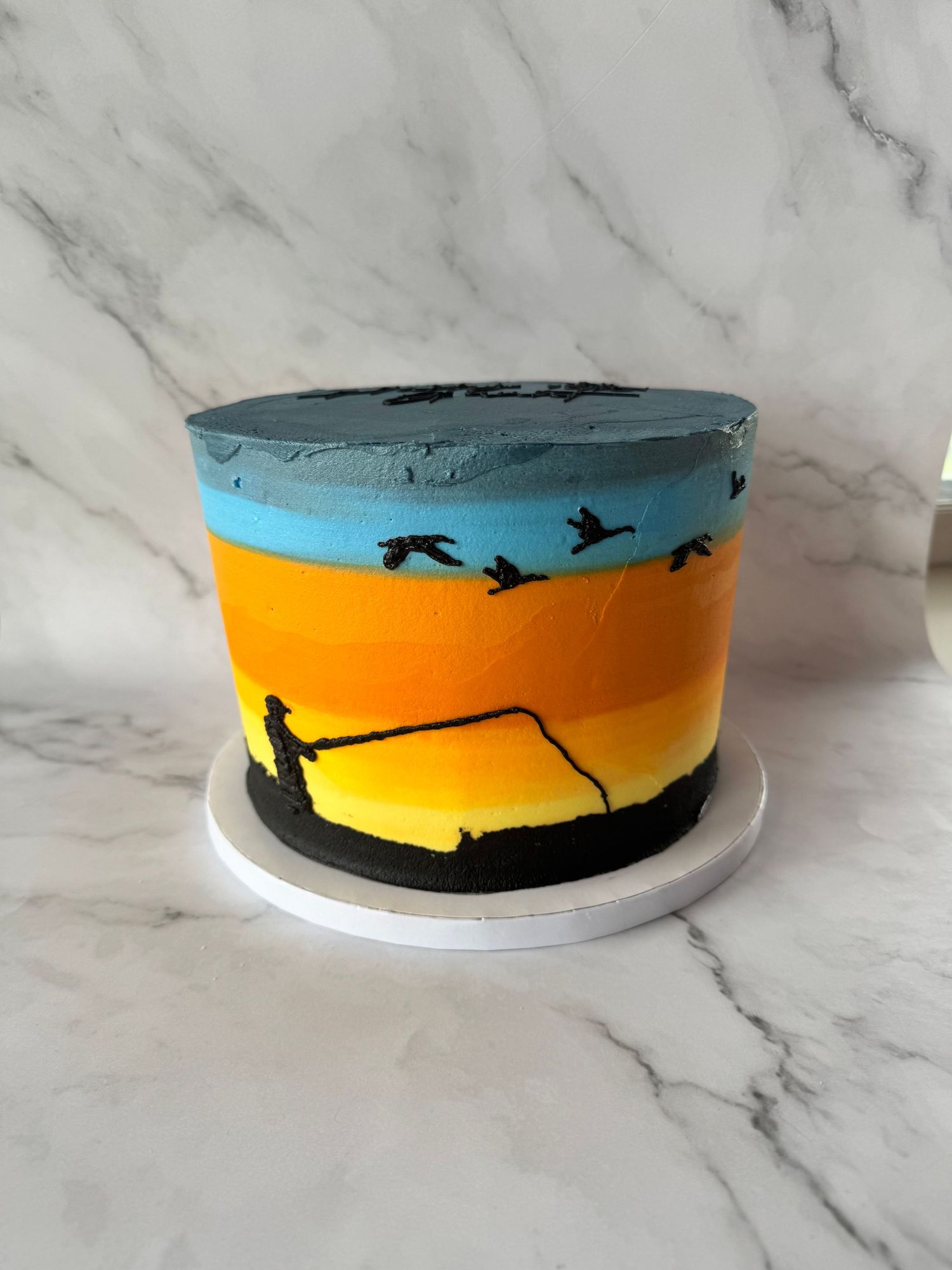 A decorative cake with a painted sunset design featuring a blue sky, orange horizon, and black silhouette details on the sides.