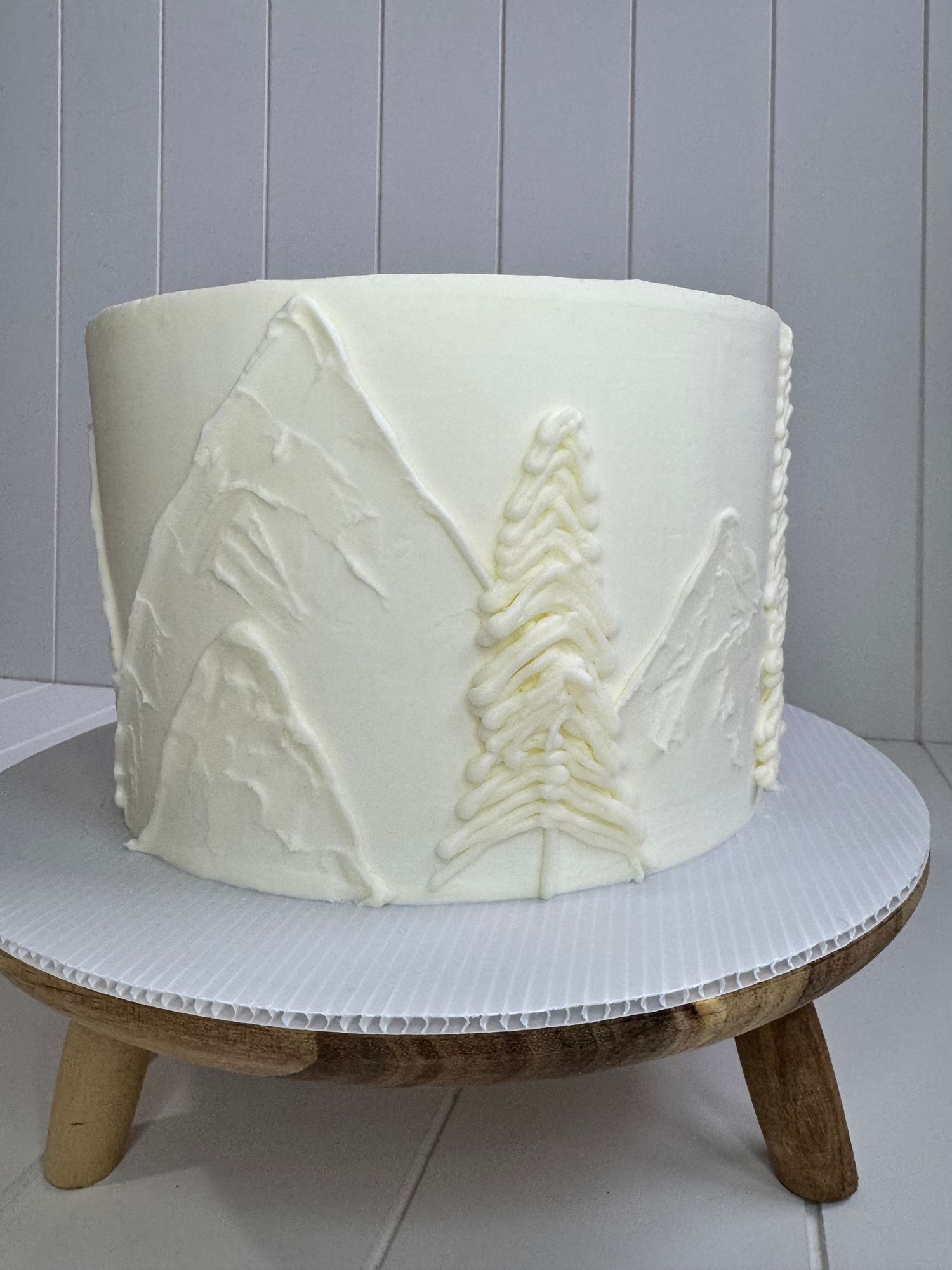 White frosted cake with delicate tree branch designs piped in buttercream on a wooden cake stand.