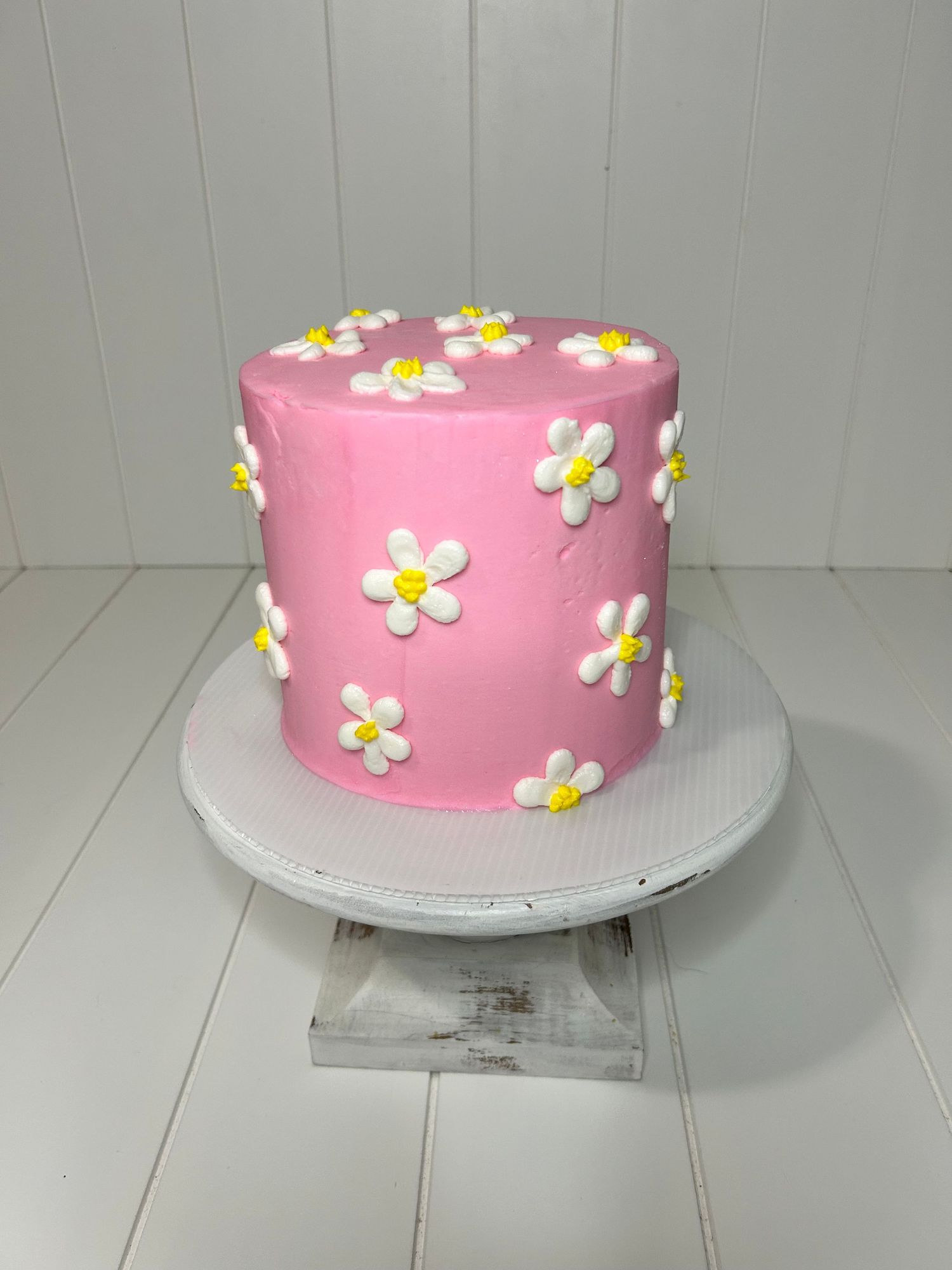 Pink birthday cake decorated with small white and yellow daisy flowers on a white stand.