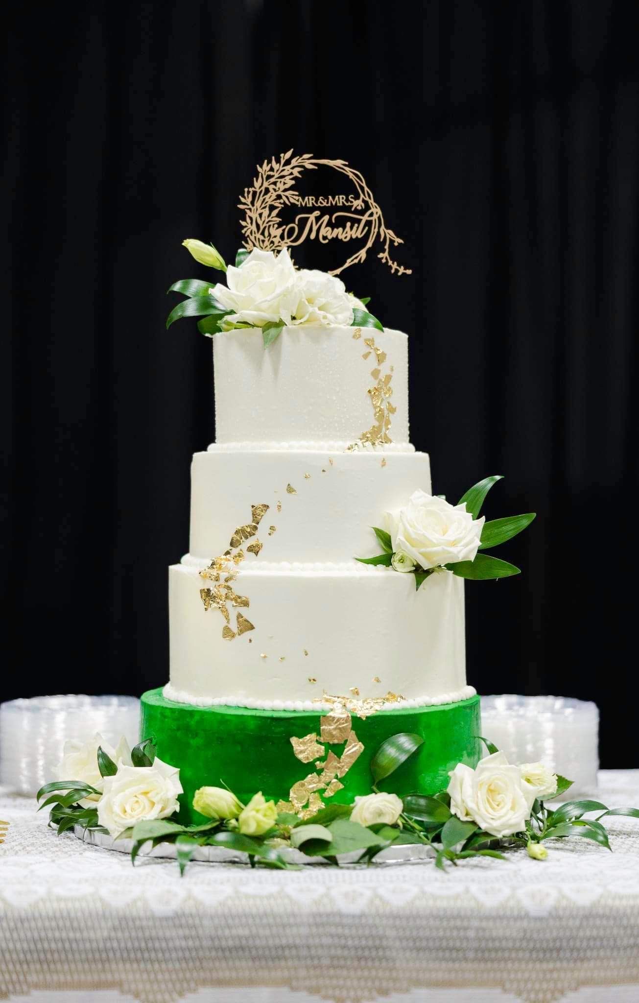 Three-tiered white wedding cake with green base and white roses accented with gold leaf details.