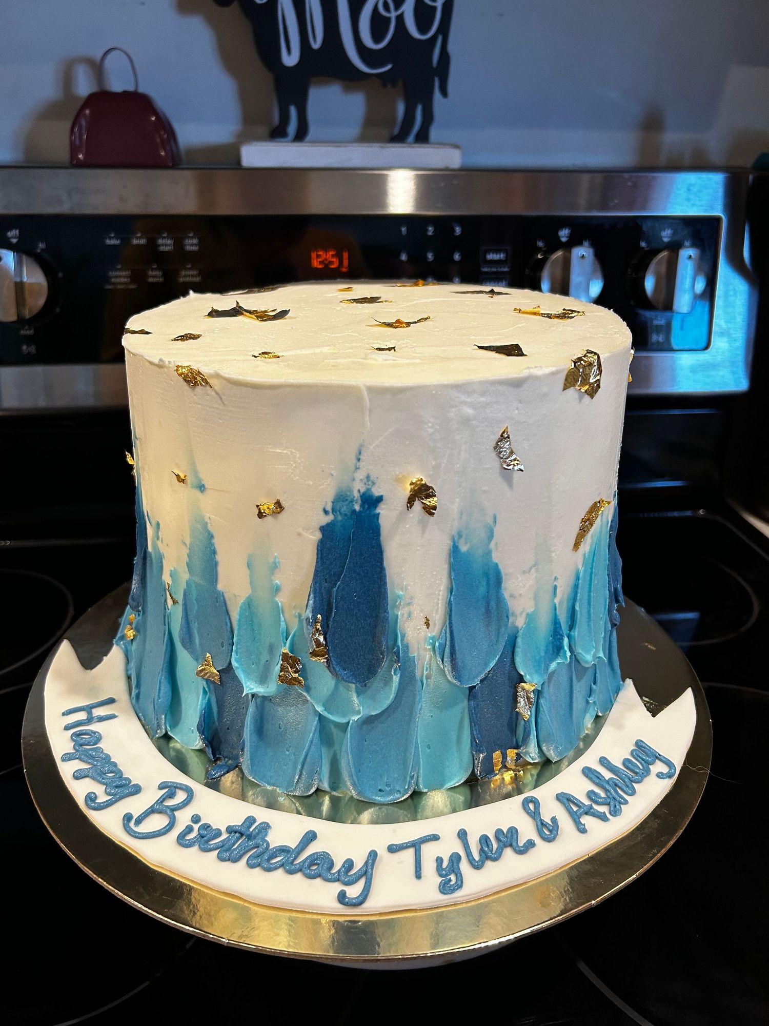 A white birthday cake with blue watercolor-style drip effect and gold accents around the sides.