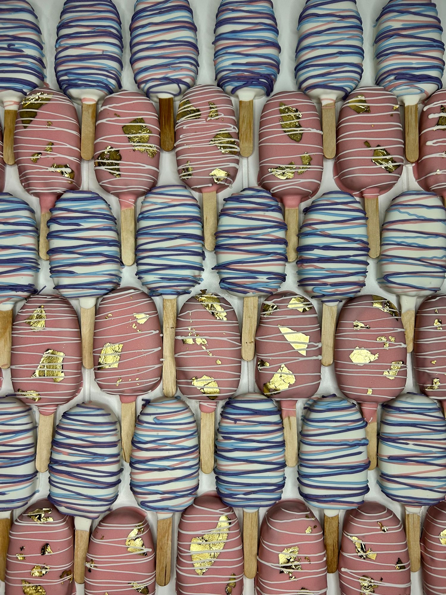 Artfully decorated cake pops with pink and blue striped designs and gold accents arranged in rows.
