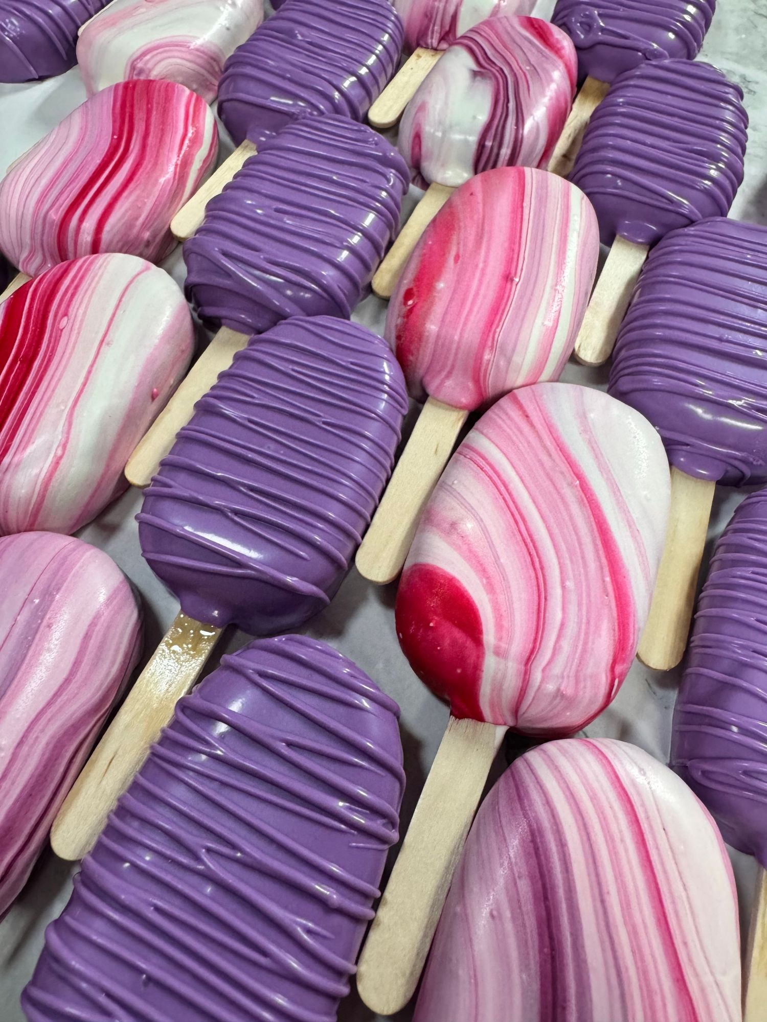 Colorful cake pops arranged in rows featuring purple and pink marbled frosting with white drizzle on wooden sticks.