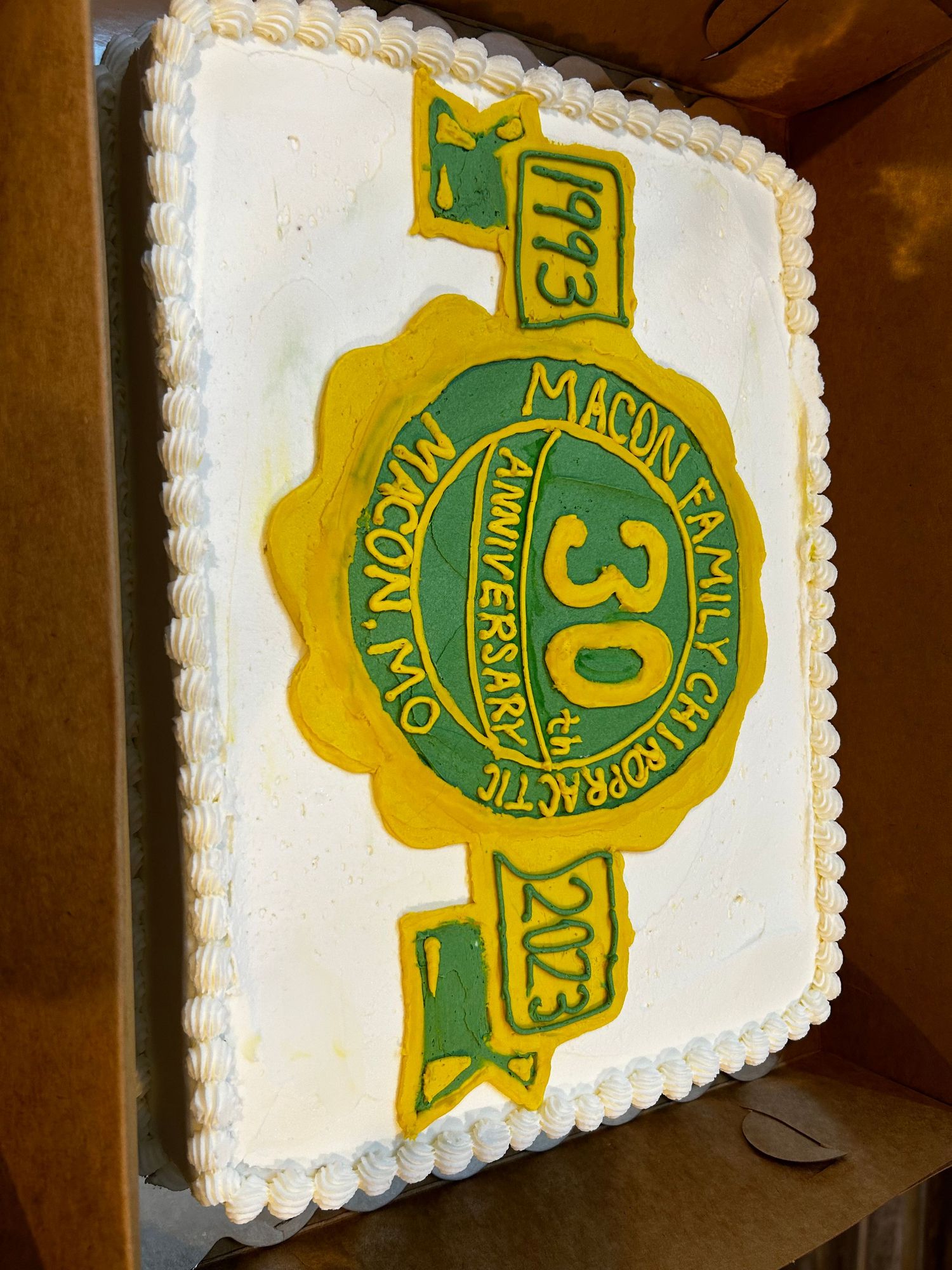 Yellow and green commemorative badge design on white square cake with decorative piped border.