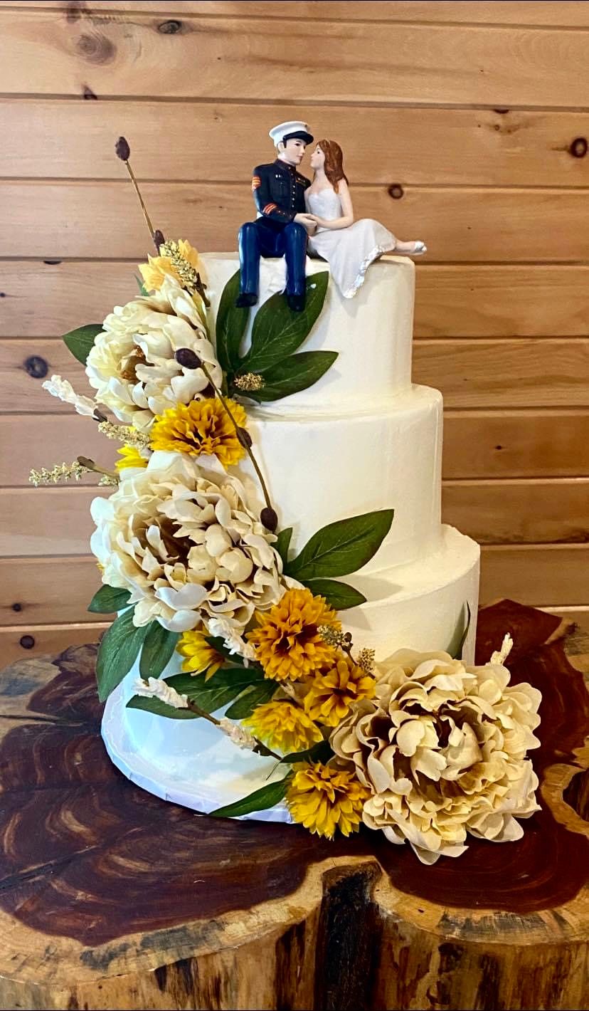 White wedding cake adorned with yellow and cream flowers on rustic wood slice stand.
