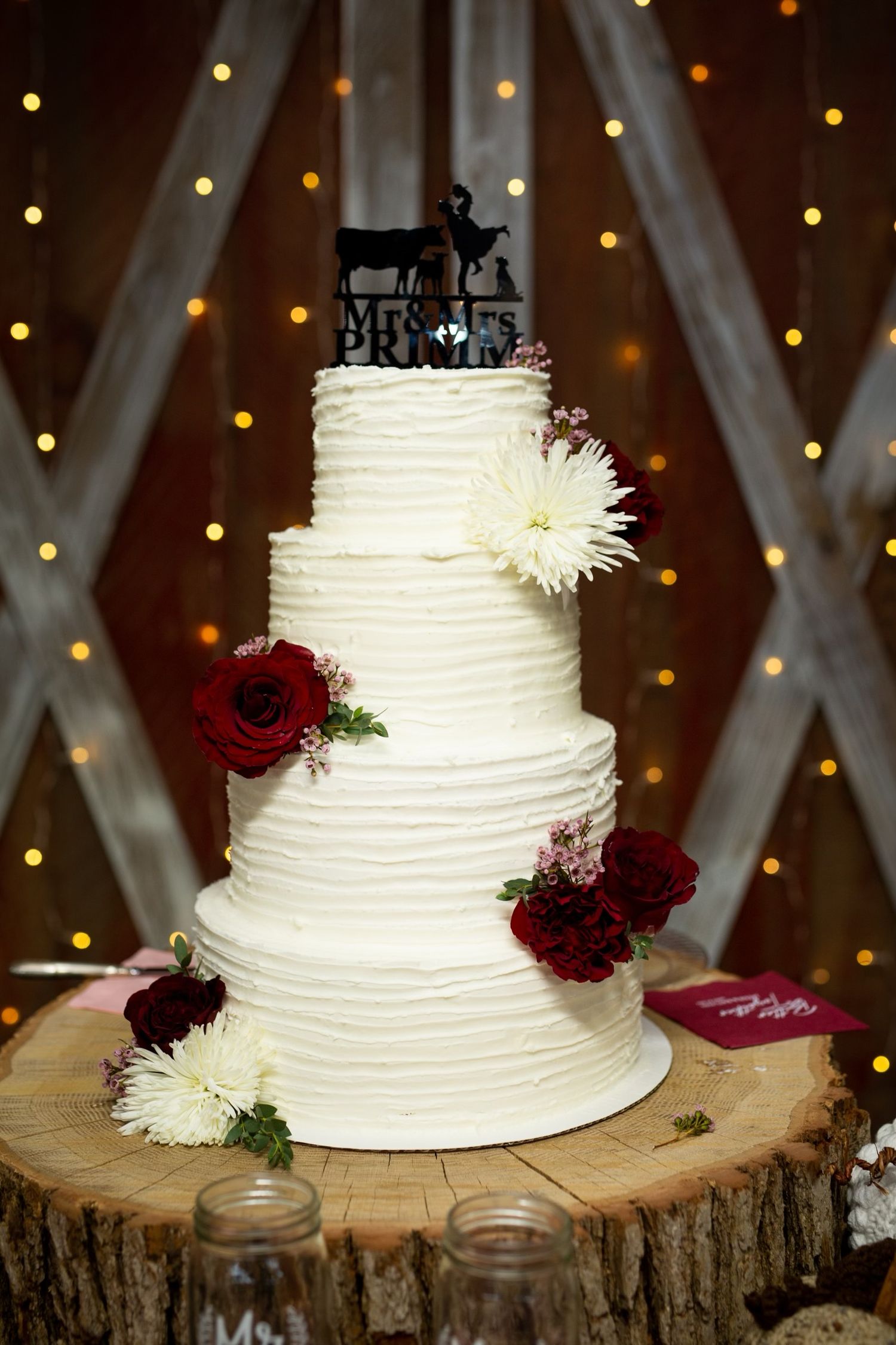 Elegant white wedding cake with burgundy flowers displayed on rustic wood slice with twinkle lights.