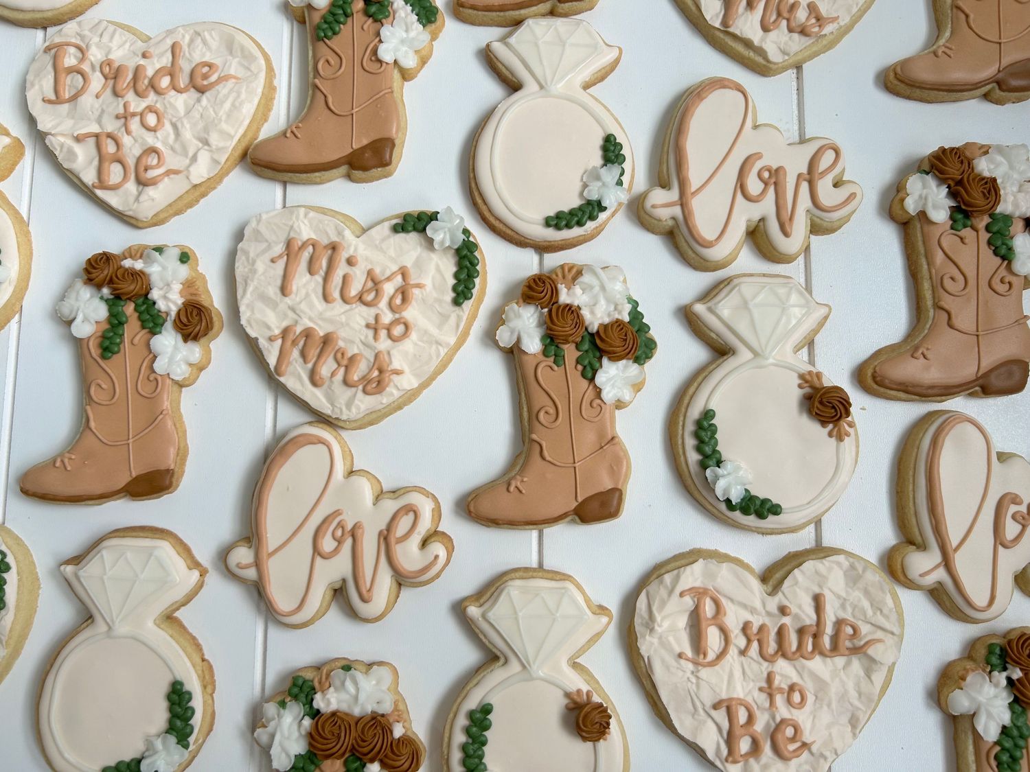 Wedding themed sugar cookies with cowboy boots, hearts, and bride-to-be text in blush pink.